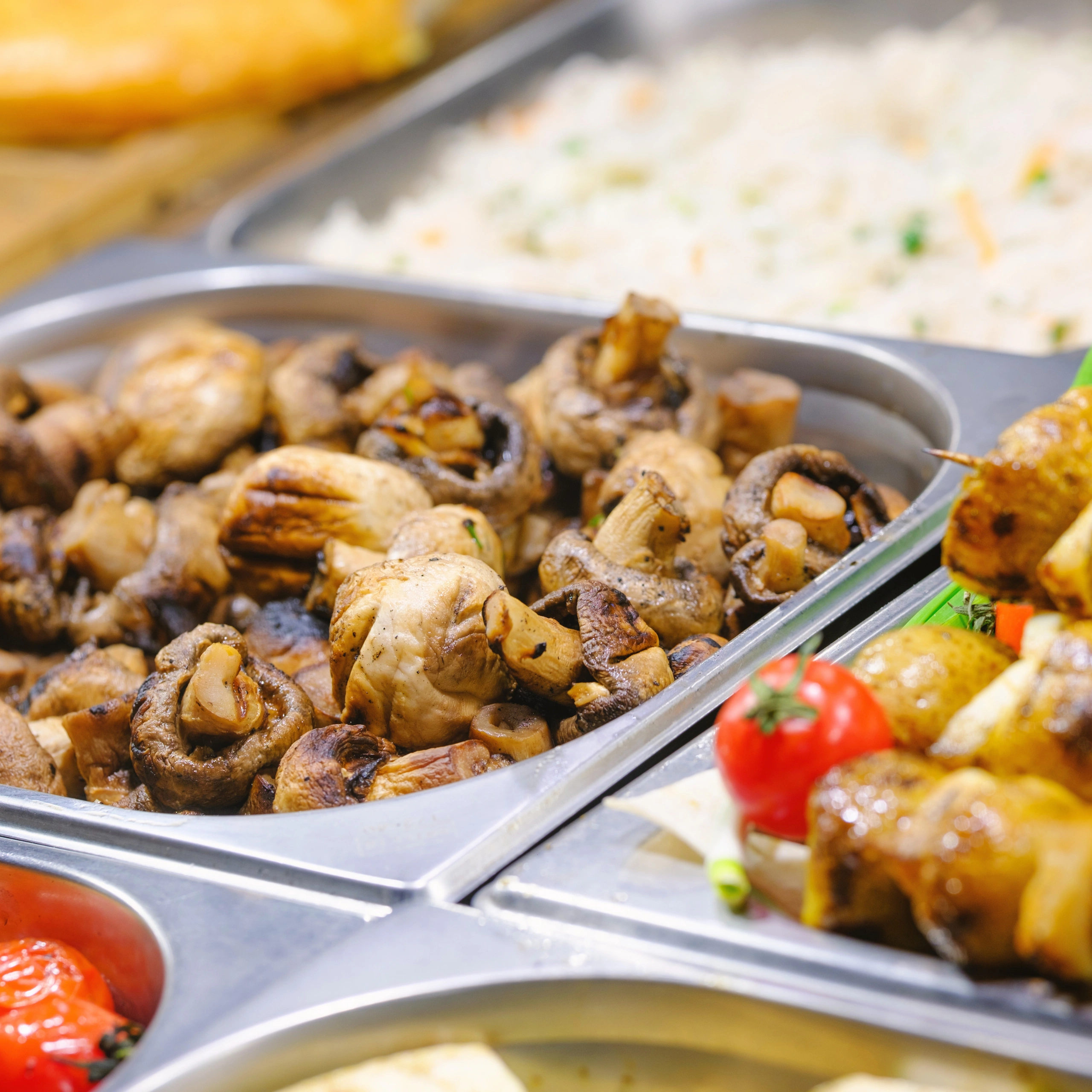 A buffet tray holds browned roasted chicken pieces and mushrooms, with creamy rice and grilled vegetables nearby, creating a warm, hearty meal display.