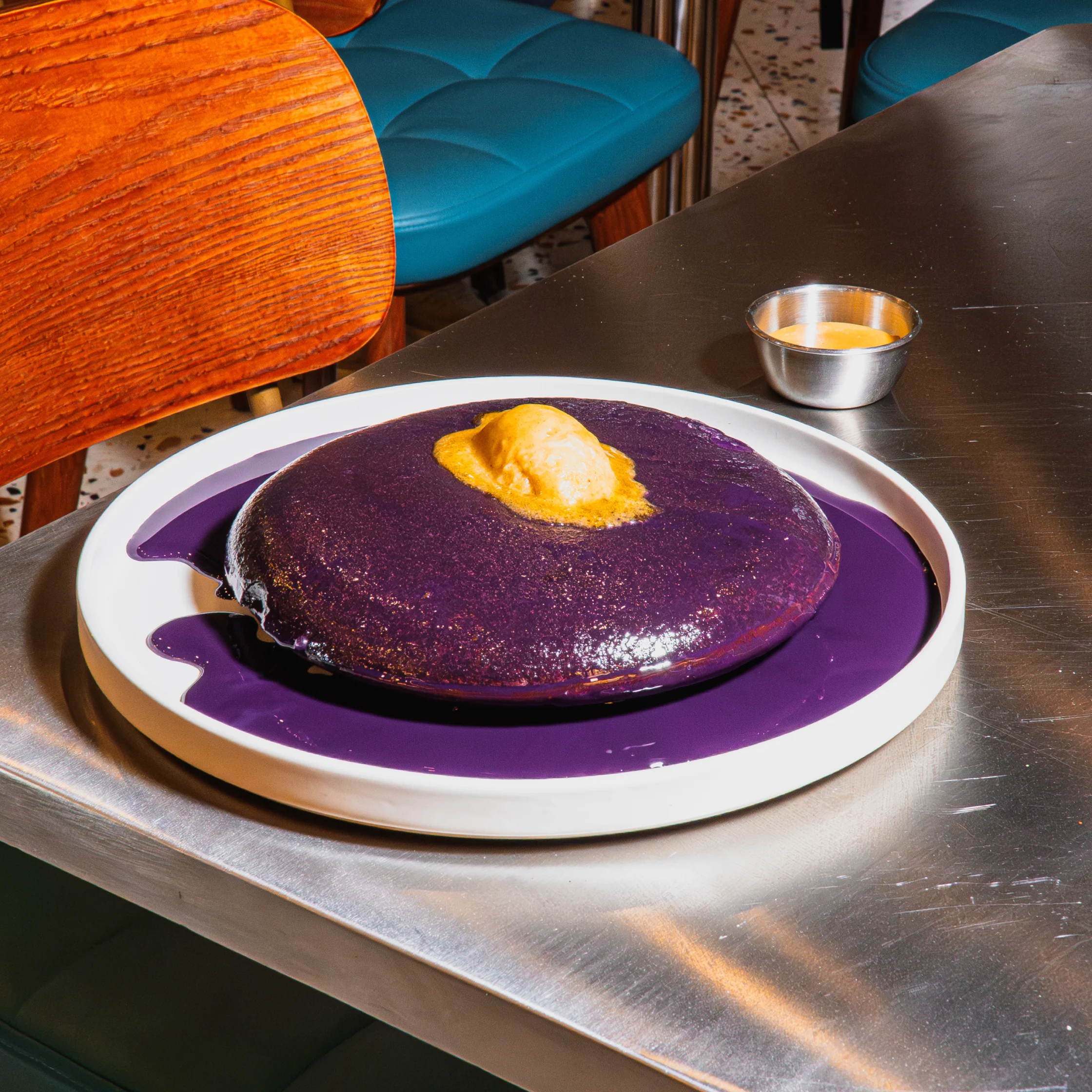 A glossy purple pancake topped with a pat of butter sits on a white plate with purple sauce, on a metal café table; vivid, playful presentation.