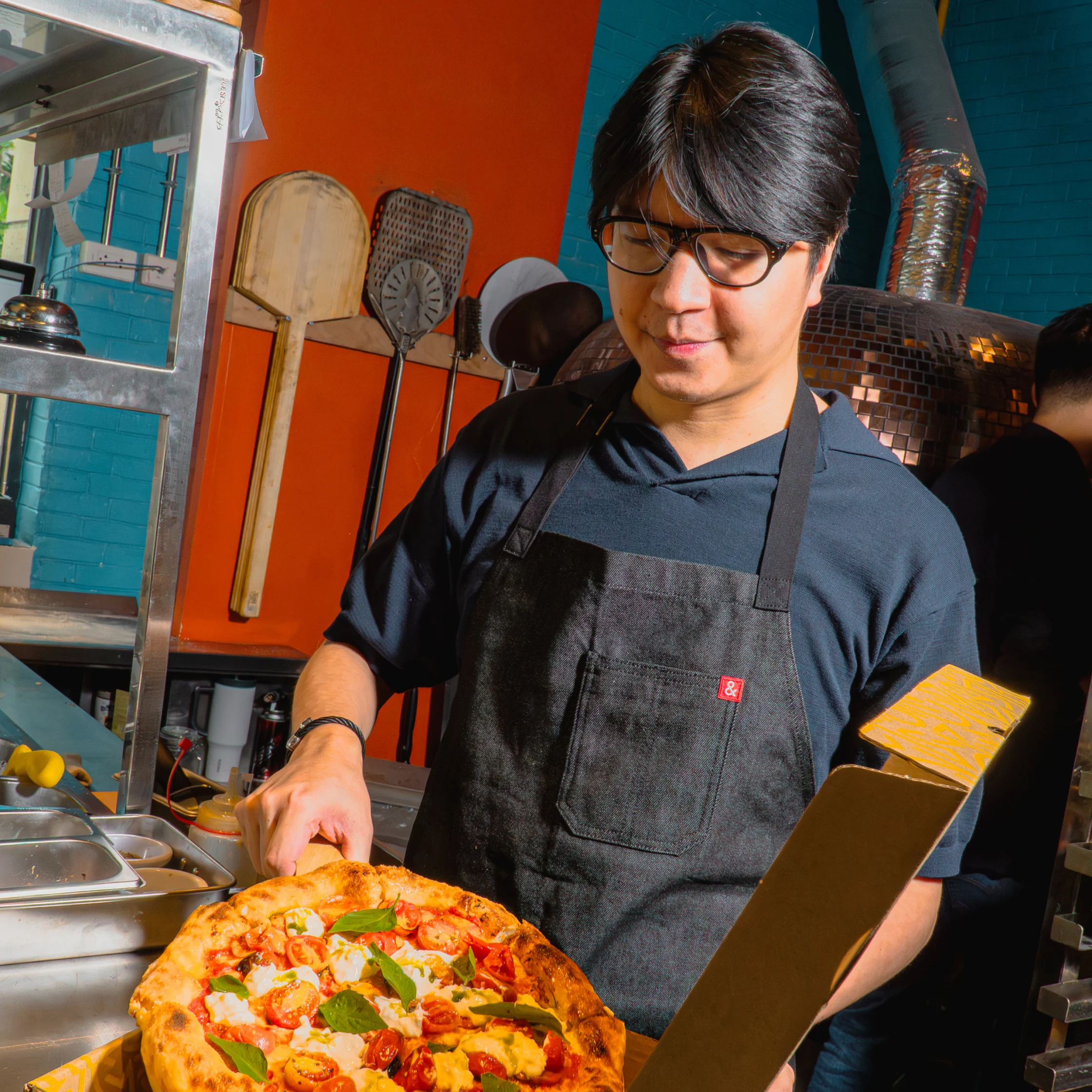 A bespectacled cook in a black apron carefully prepares a colorful, basil-topped pizza at a busy restaurant counter, focused and calm.