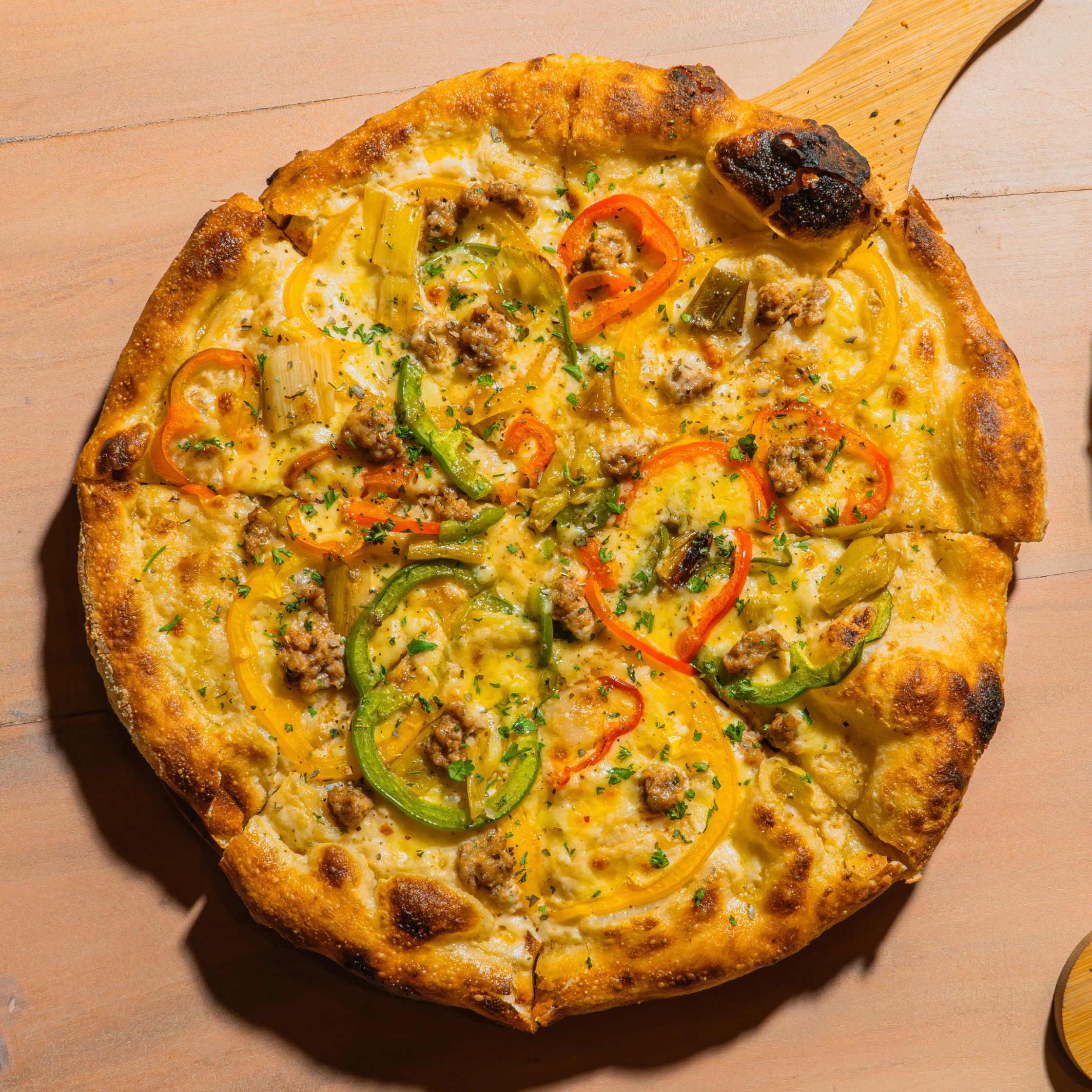Golden-crusted vegetable pizza topped with melted cheese, tomato, green pepper, and herbs on a wooden board, with a wooden serving spatula; warm, appetizing scene.