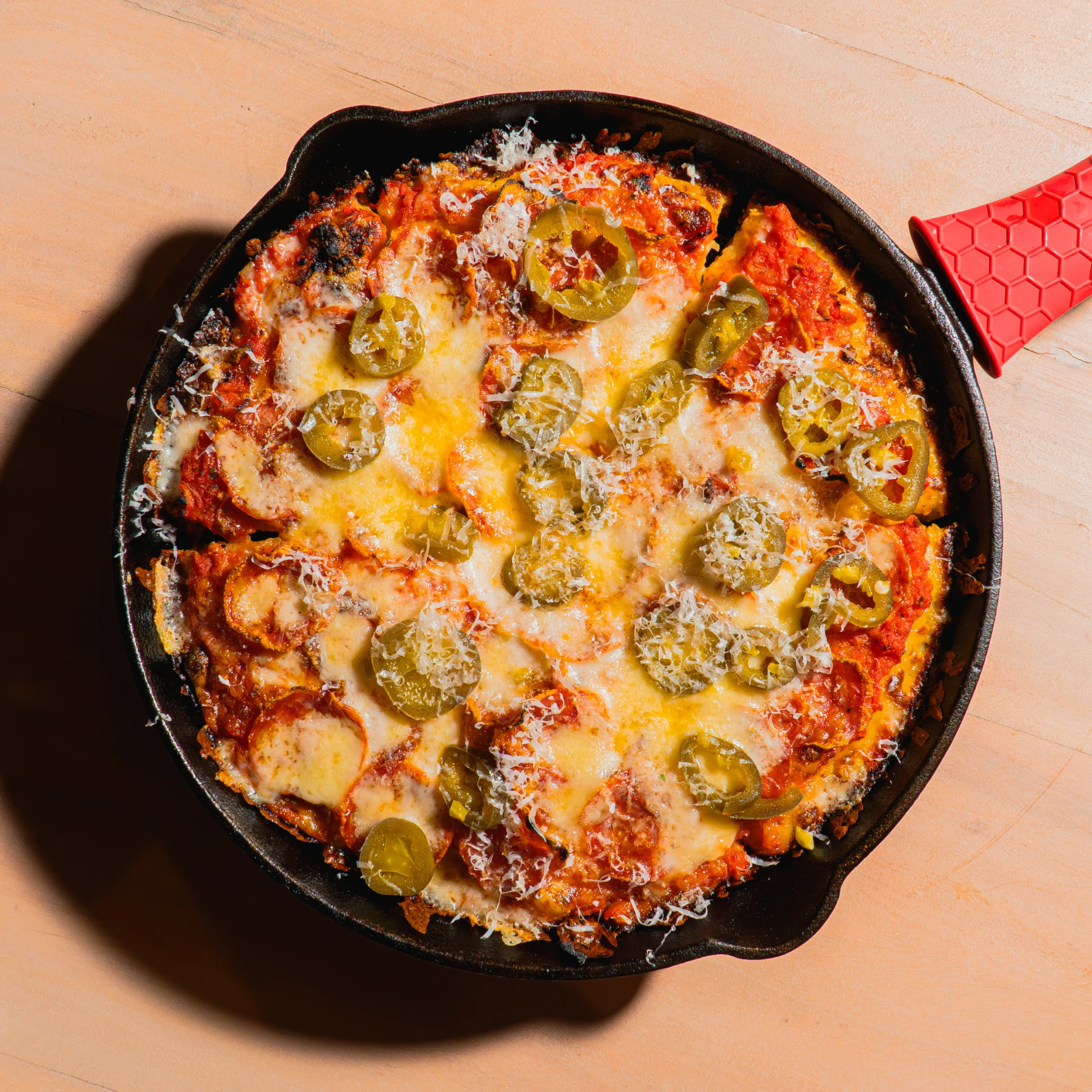 Overhead view of a bubbling, cheesy pizza topped with pepperoni and jalapeños in a black cast-iron skillet, with a red handle on a wooden surface.