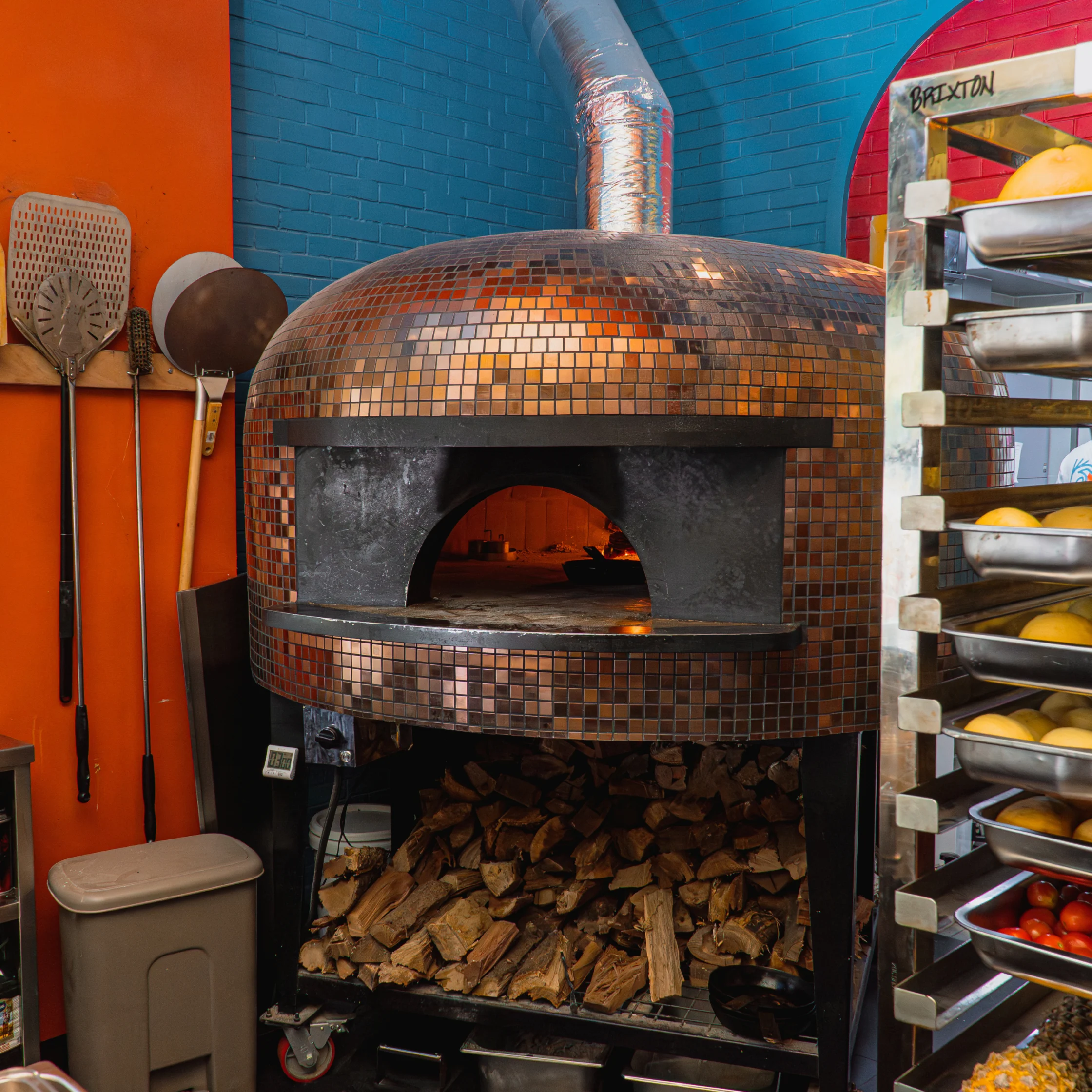 Copper-tiled wood-fired pizza oven glows against blue and orange brick walls, with stacked firewood, pizza paddles, and ingredient trays nearby.