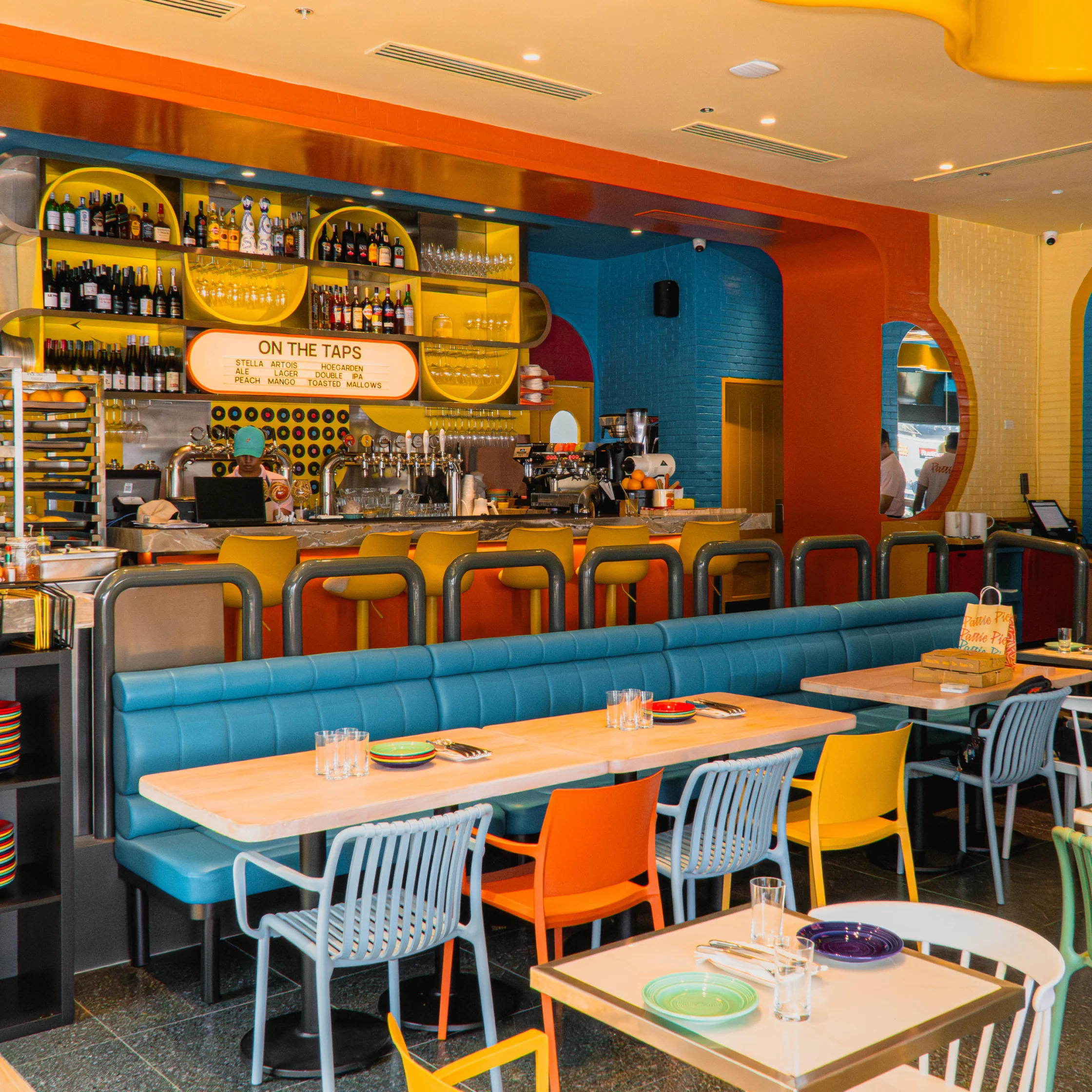 A vibrant modern restaurant and bar with a turquoise booth, colorful orange and yellow chairs, a stocked bottle-lined bar, and bright, playful décor.