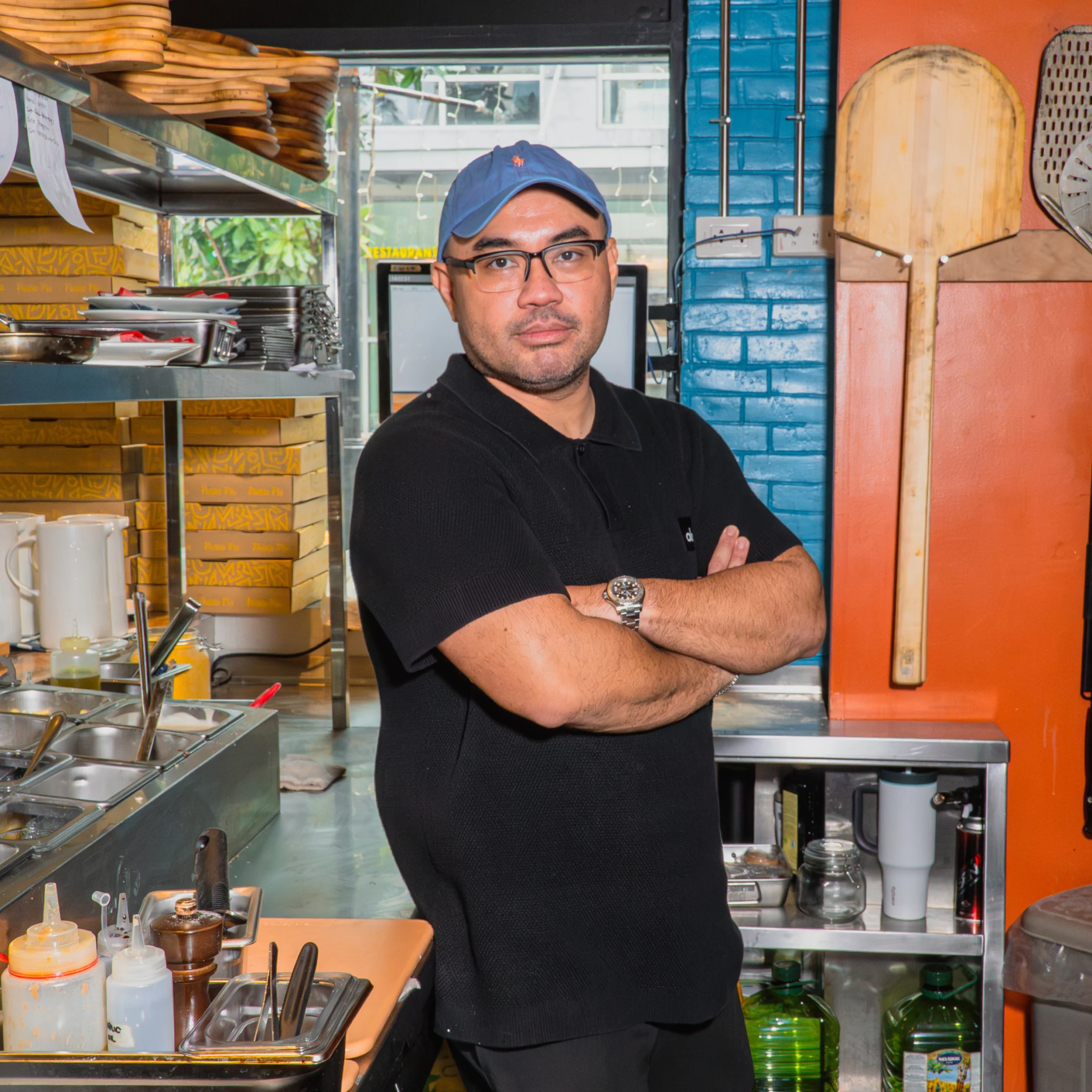A restaurant worker in a blue cap and black shirt stands with arms crossed in a busy kitchen, surrounded by cooking tools and supplies; his pose feels confident.