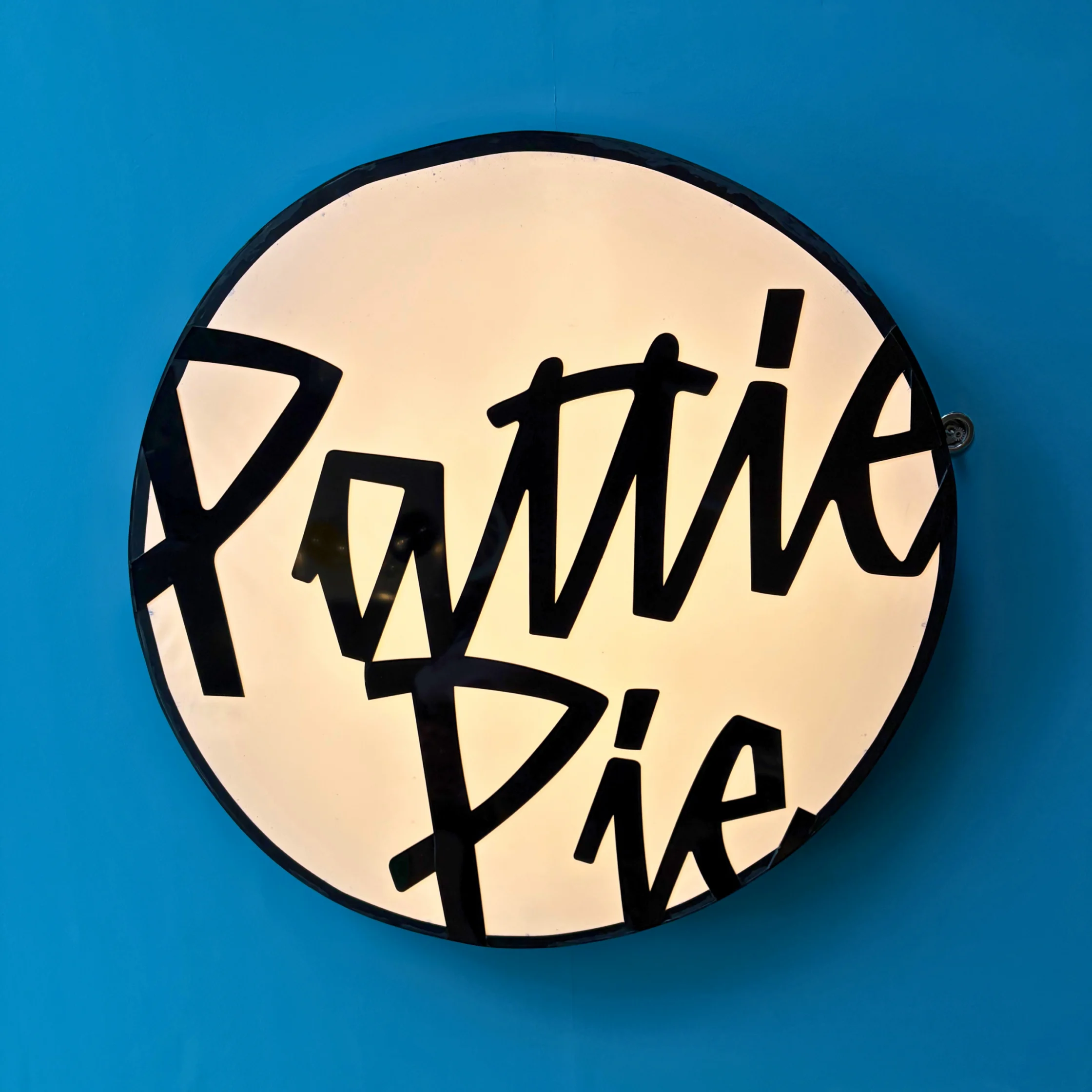 A round, cream-lit sign on a bright blue wall reads “Pattie Pie” in bold, playful black lettering, giving the handmade display a cheerful feel.