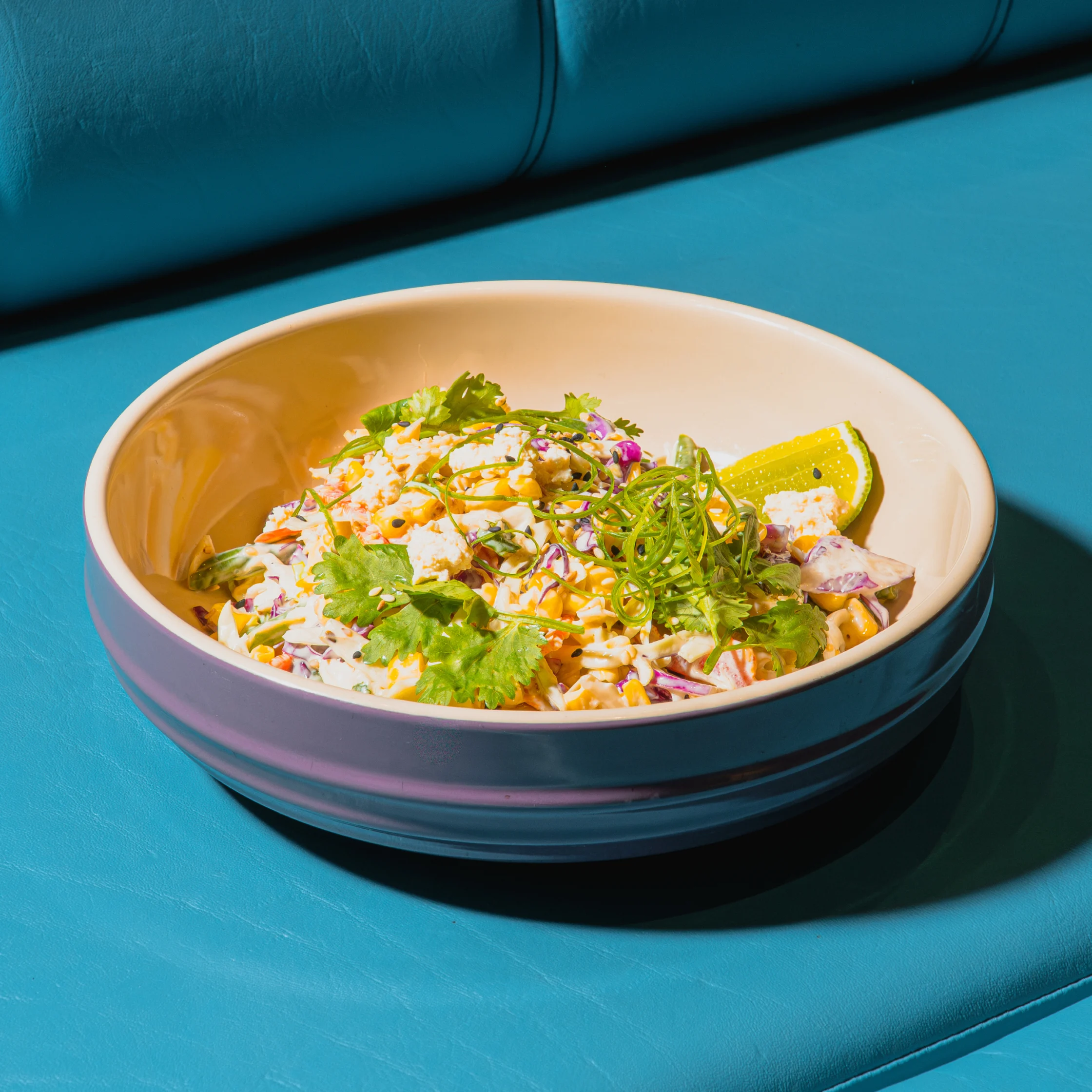 A creamy seafood salad topped with cilantro and herbs sits in a ceramic bowl with a lime wedge, against a vivid turquoise backdrop. It feels fresh and vibrant.