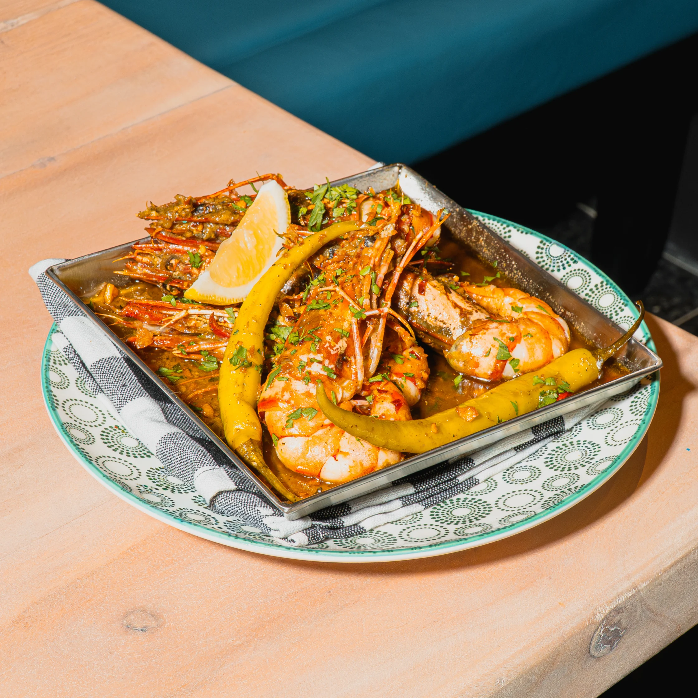 A metal tray of spicy grilled prawns with herbs, lemon, and green peppers sits on a patterned plate on a wooden table, looking vibrant and appetizing.
