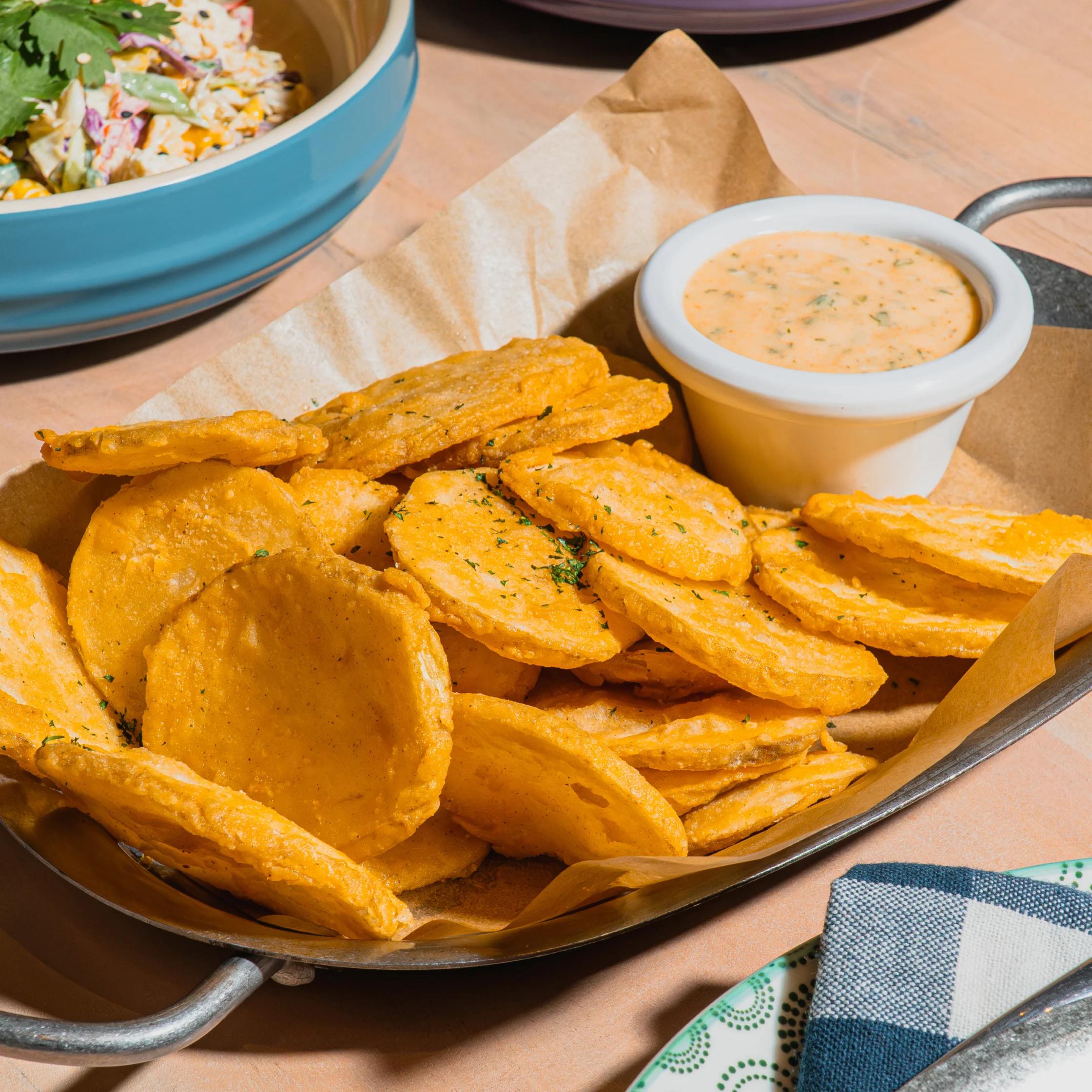 Golden seasoned potato wedges in a parchment-lined metal tray with creamy dipping sauce, beside a bowl of slaw; a warm, appetizing meal.