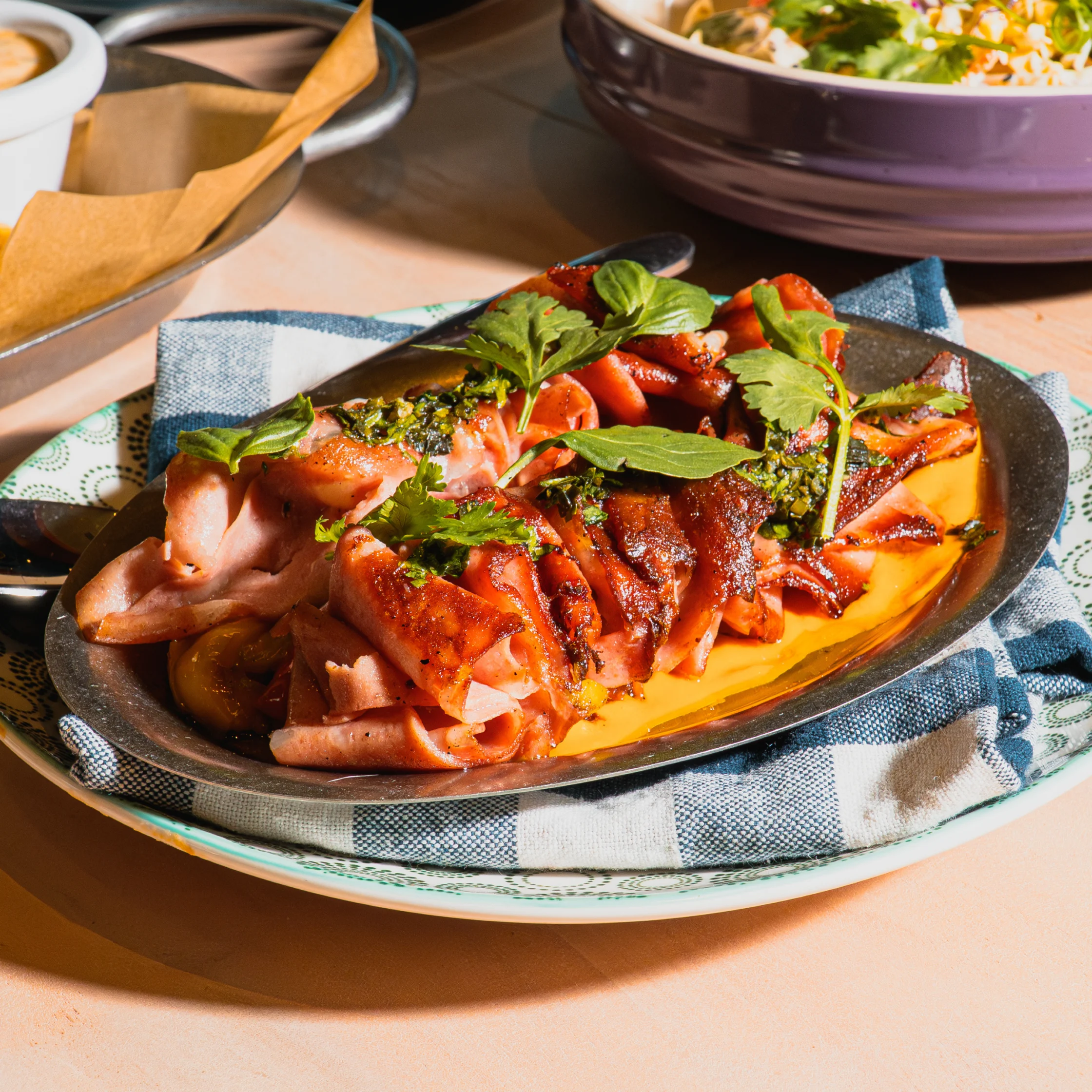 A sizzling platter of grilled sausage and ham in glossy orange sauce, topped with herbs and served on a blue-checkered cloth; a warm, appetizing meal.