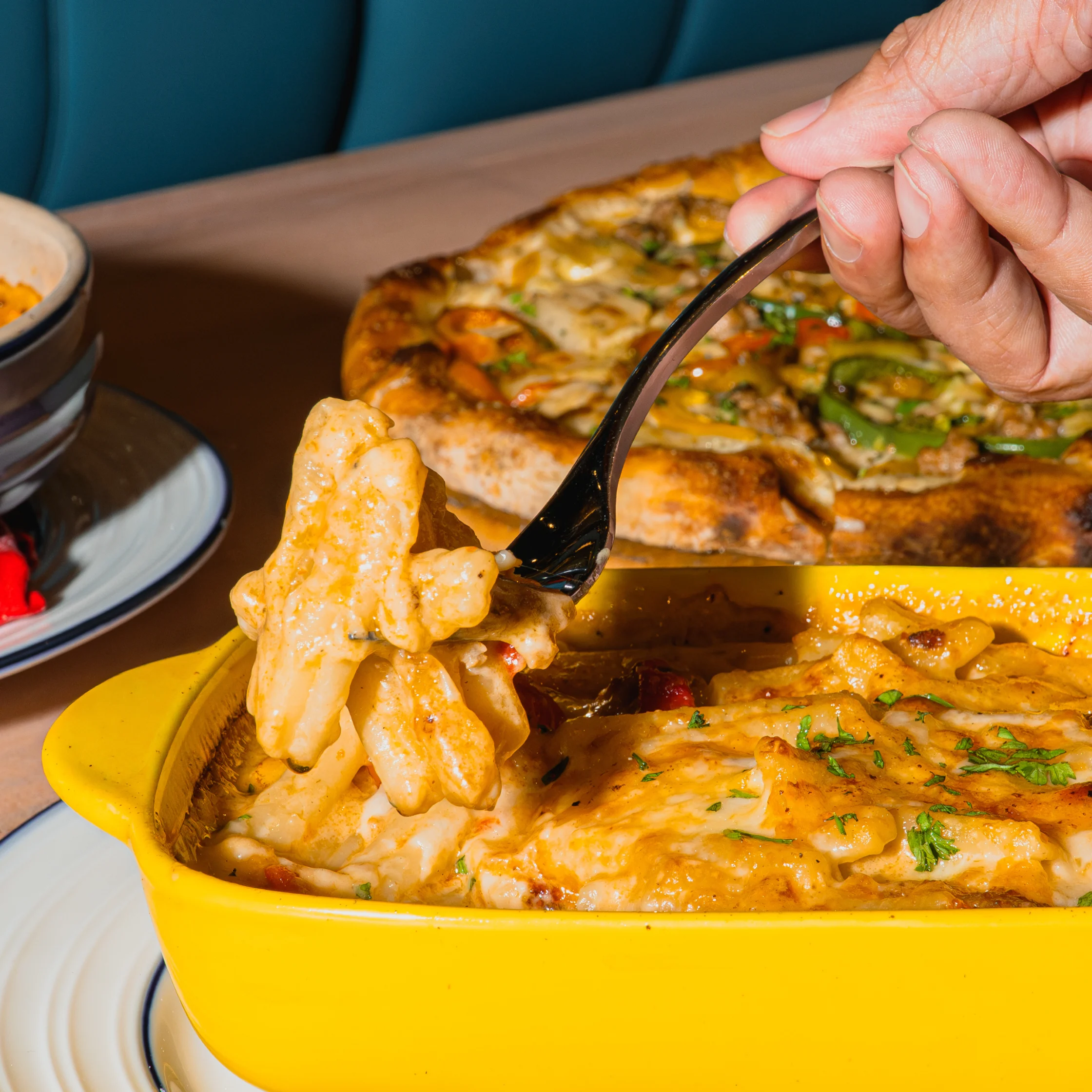 A hand lifts creamy baked pasta from a bright yellow casserole dish, with a golden vegetable-topped pizza in the background; warm, inviting restaurant meal.