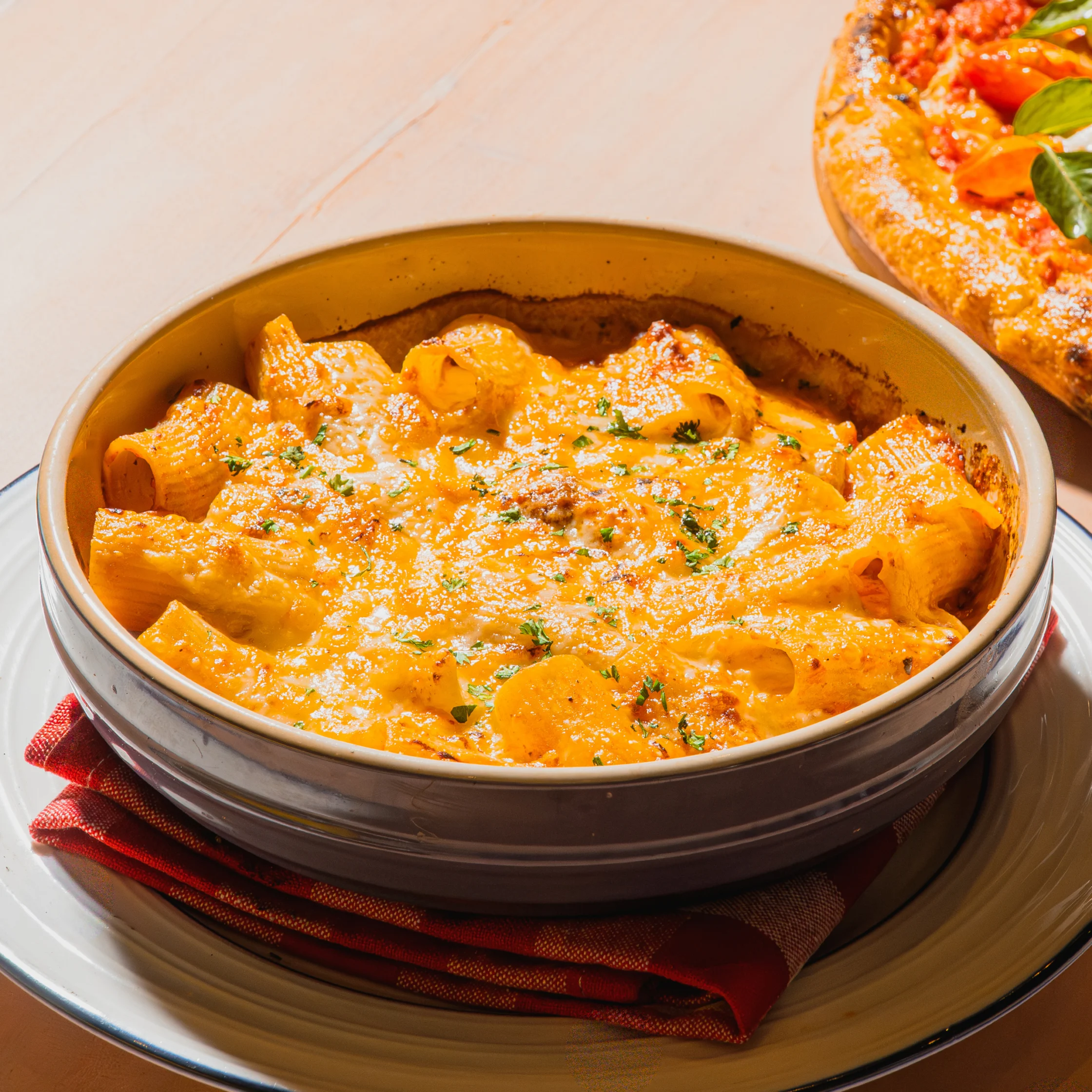 A creamy orange curry with paneer chunks and herbs fills a ceramic bowl on a red napkin; a tomato-topped dish peeks in, creating a warm, appetizing scene.