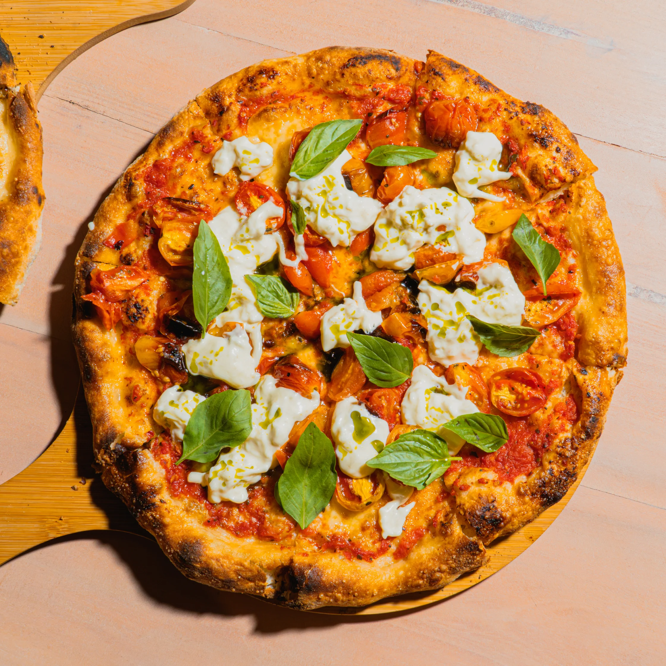 Overhead view of a golden, charred-crust pizza topped with tomato sauce, fresh basil, cherry tomatoes, and creamy mozzarella in warm sunlight.