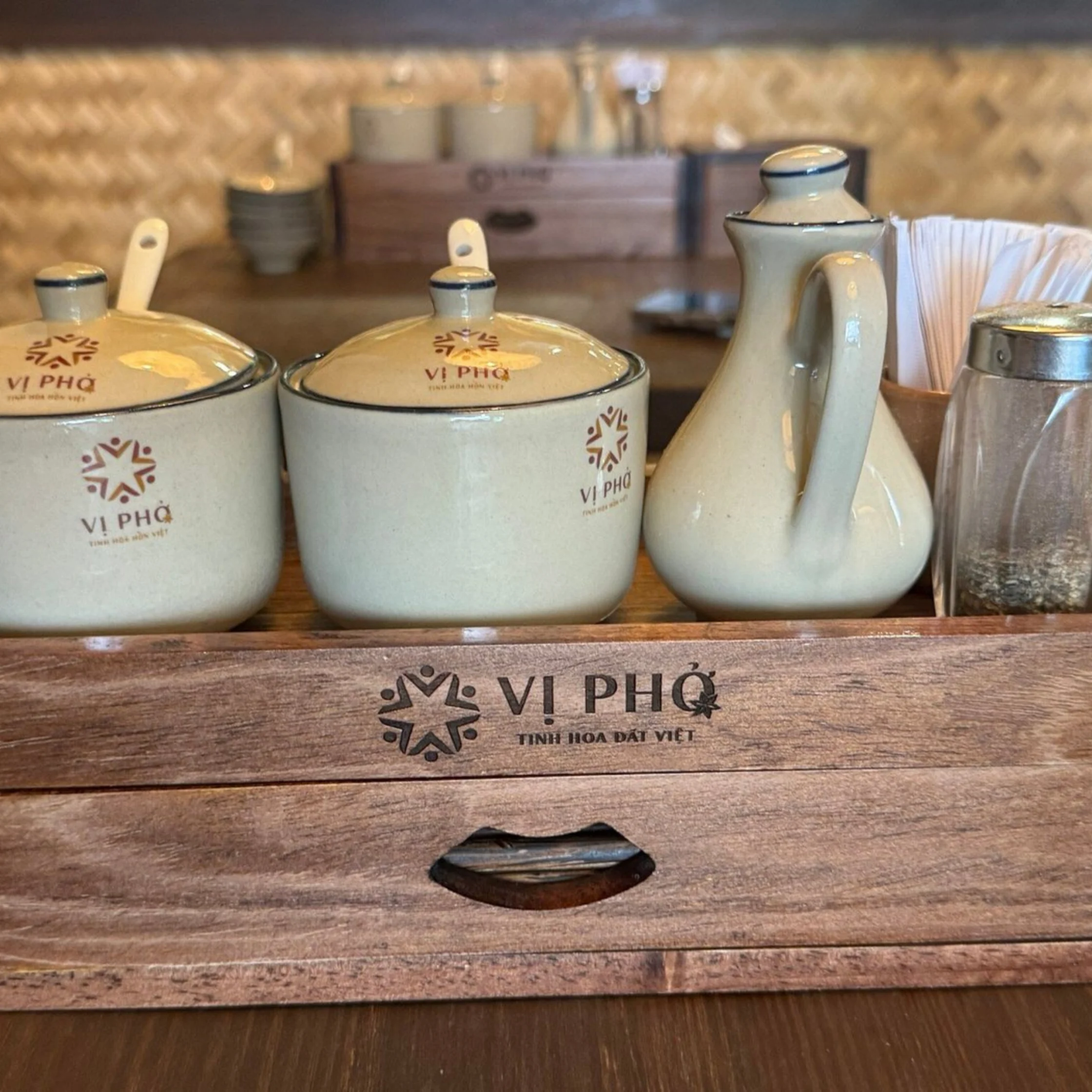 Cream-colored ceramic tea jars, a tall pitcher and a glass of tea sit in a wooden tray, branded “Vi Phở”; warm lighting creates a cozy, traditional mood.