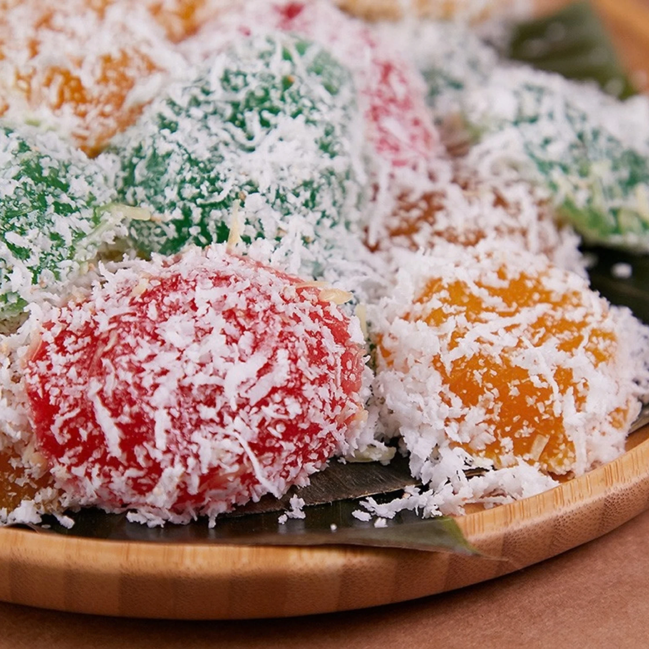 Close-up of colorful pichi-pichi sweets—red, green, orange, and pink cubes—coated in shredded coconut on a wooden platter, festive and inviting.