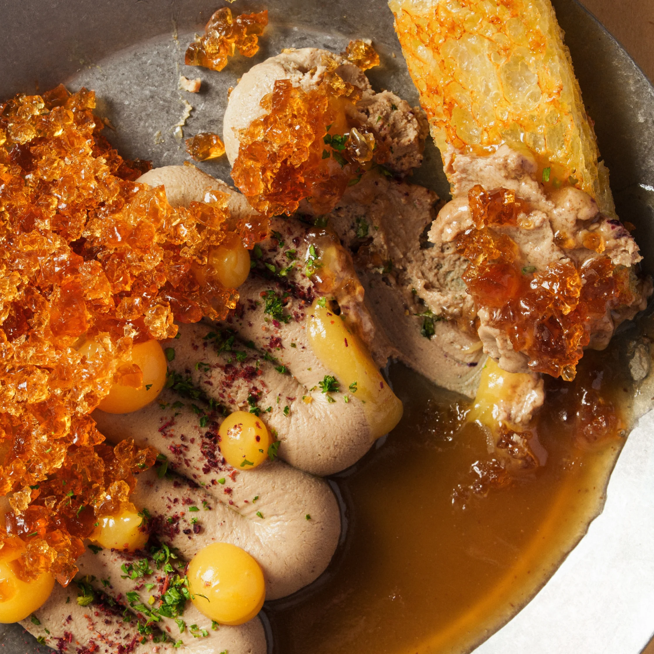 Close-up of a rich savory dish with smooth pâté, glossy brown broth, golden egg yolks, crisp amber shards, herbs, and toasted bread; indulgent and refined.
