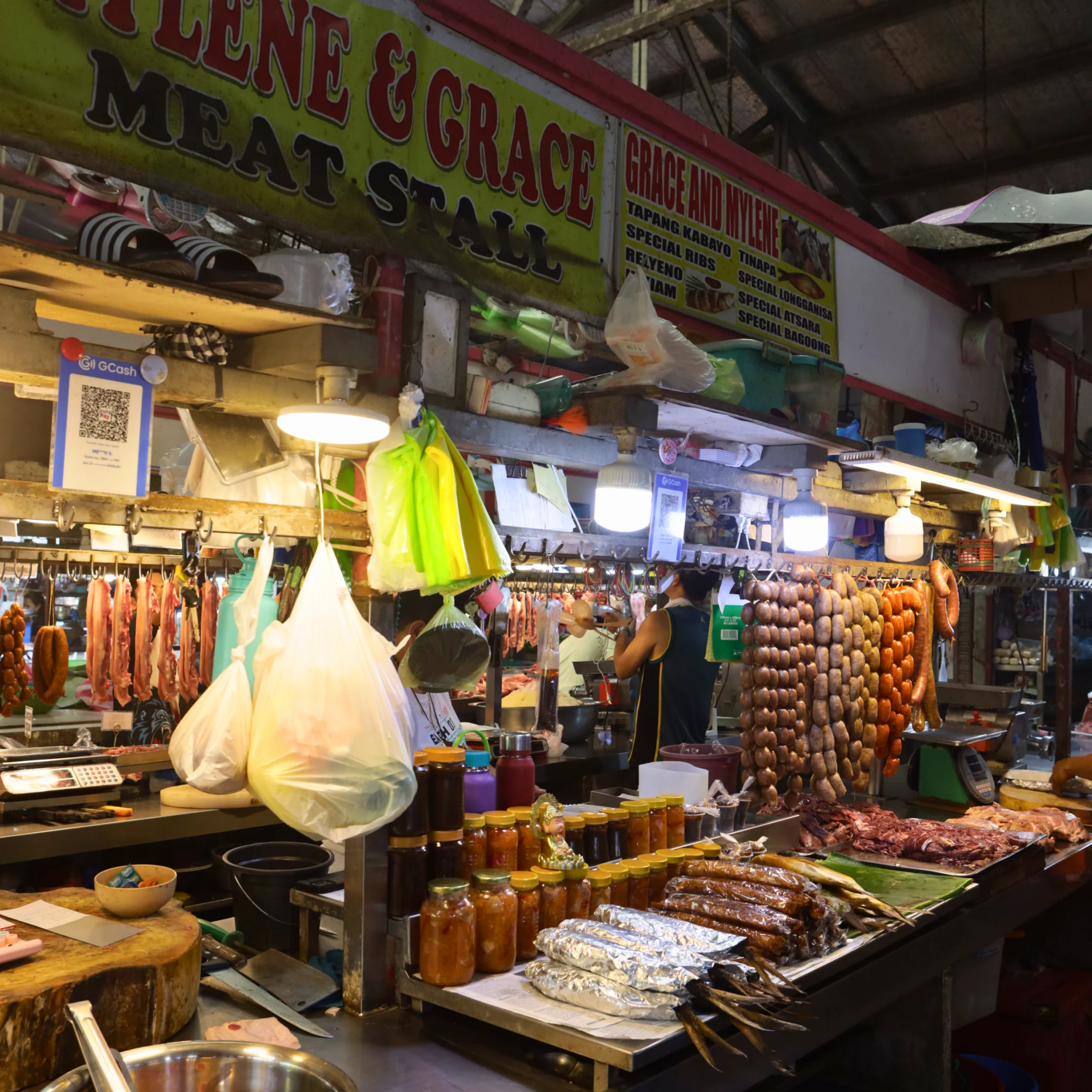 A brightly lit market stall labeled “Grace and Merle Meat Stall” displays hanging sausages, cured meats, jars, and packaged goods, creating a bustling, lively atmosphere.