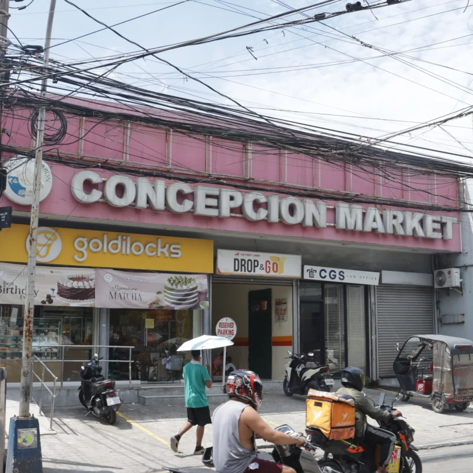 A busy street view of the pink Concepcion Market storefront, with shops and parked motorcycles below a web of overhead wires; the scene feels bustling and urban.