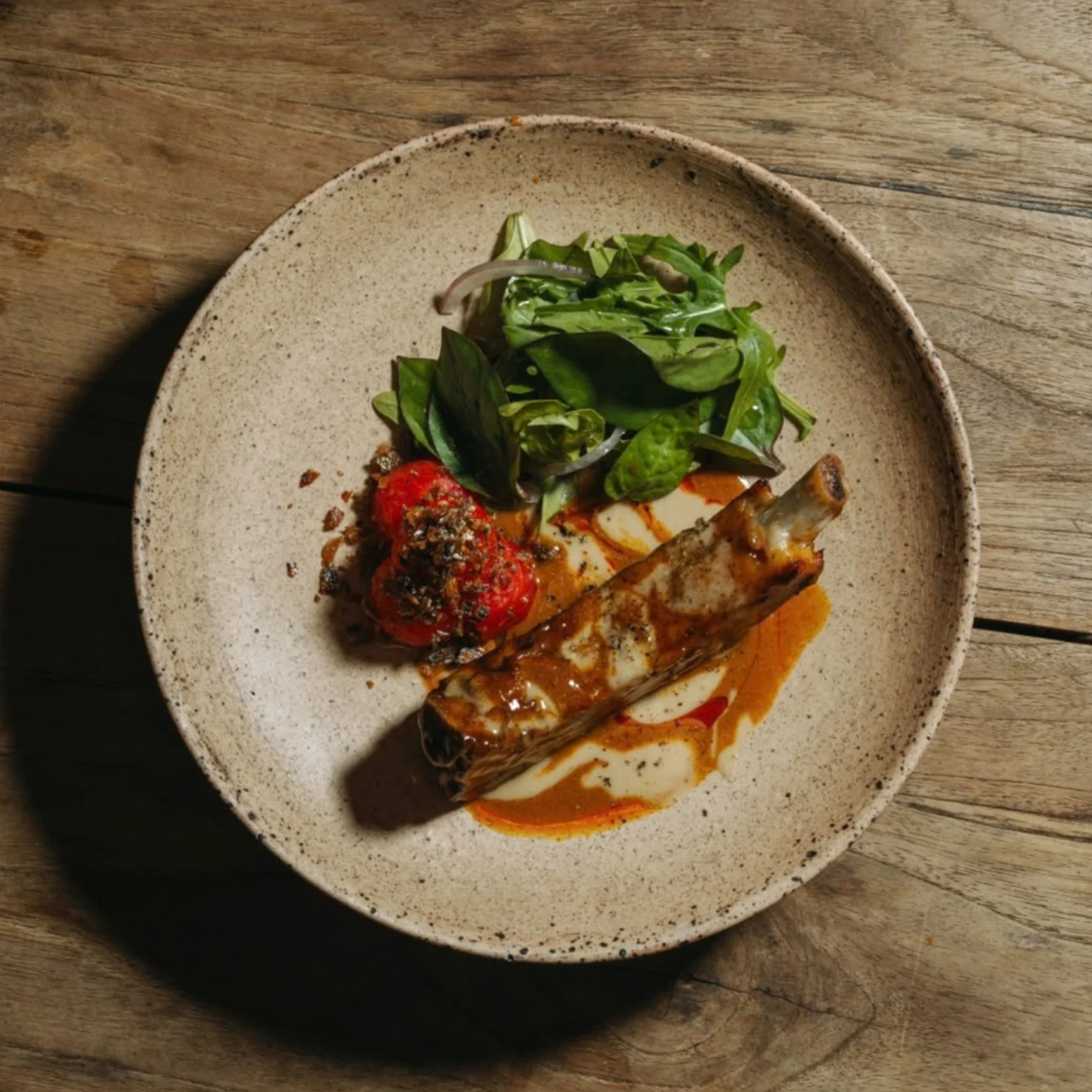 A roasted bone-in meat cut with red tomato relish and fresh greens, drizzled with sauce on a rustic ceramic plate atop a wooden table.
