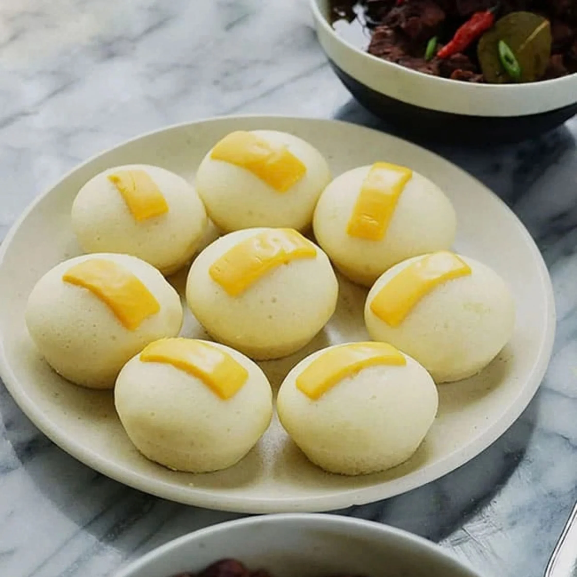 Eight round steamed buns topped with strips of yellow cheese sit on a white plate beside a bowl of dark meat curry with red and green chilies, on marble.