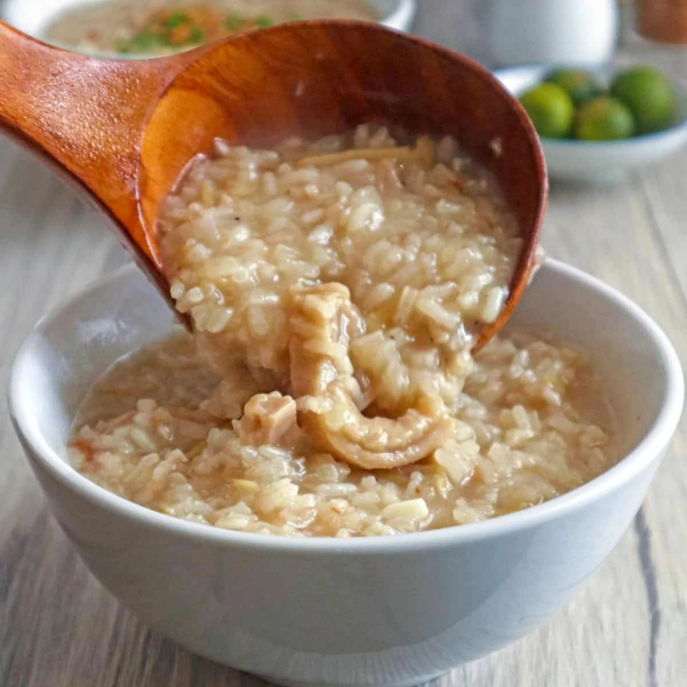 A wooden ladle pours steaming rice porridge with tender chicken into a white bowl, creating a warm, comforting meal on a bright kitchen table.