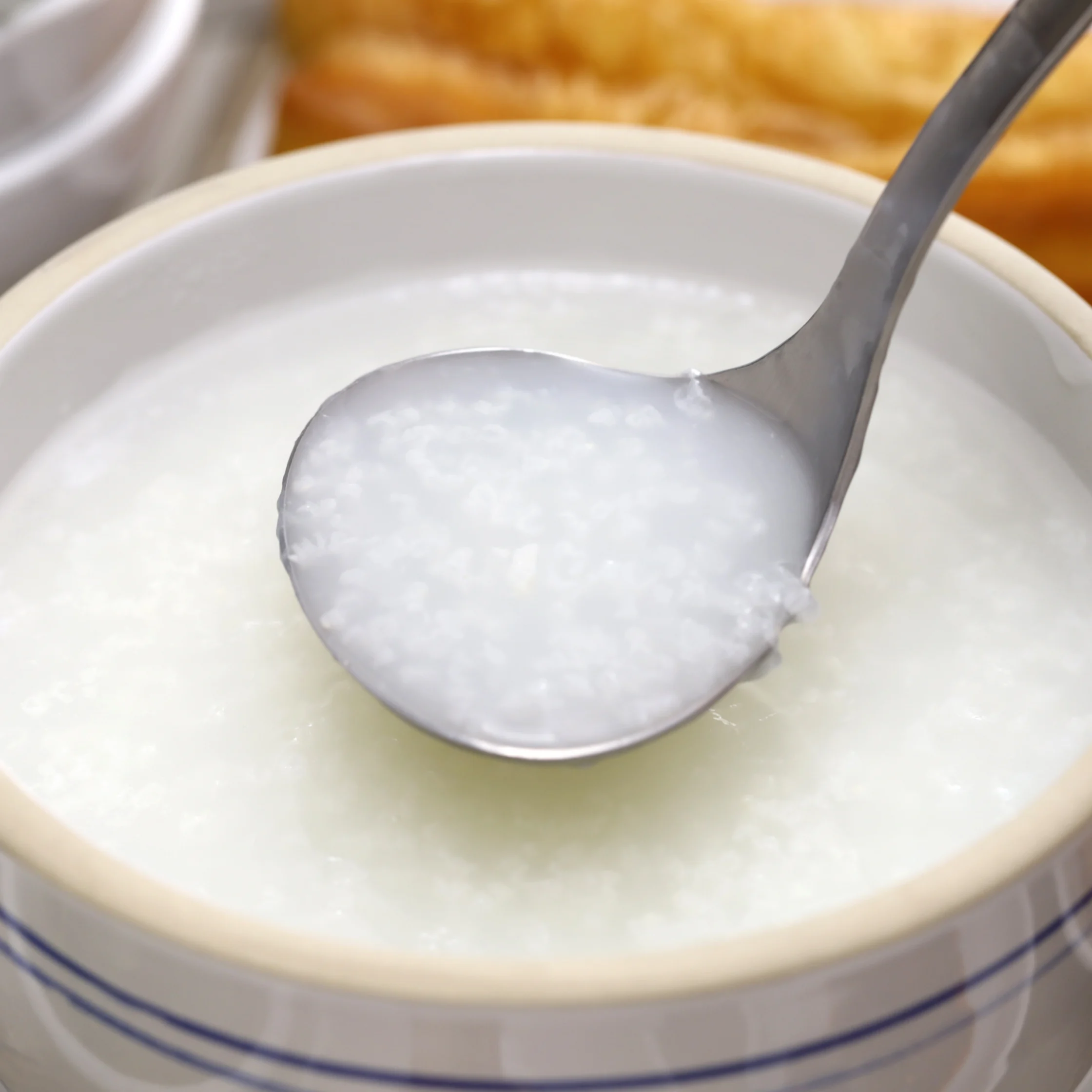 A metal spoon lifts thick white rice porridge from a ceramic bowl, with golden fried dough in the background; the scene feels warm and comforting.