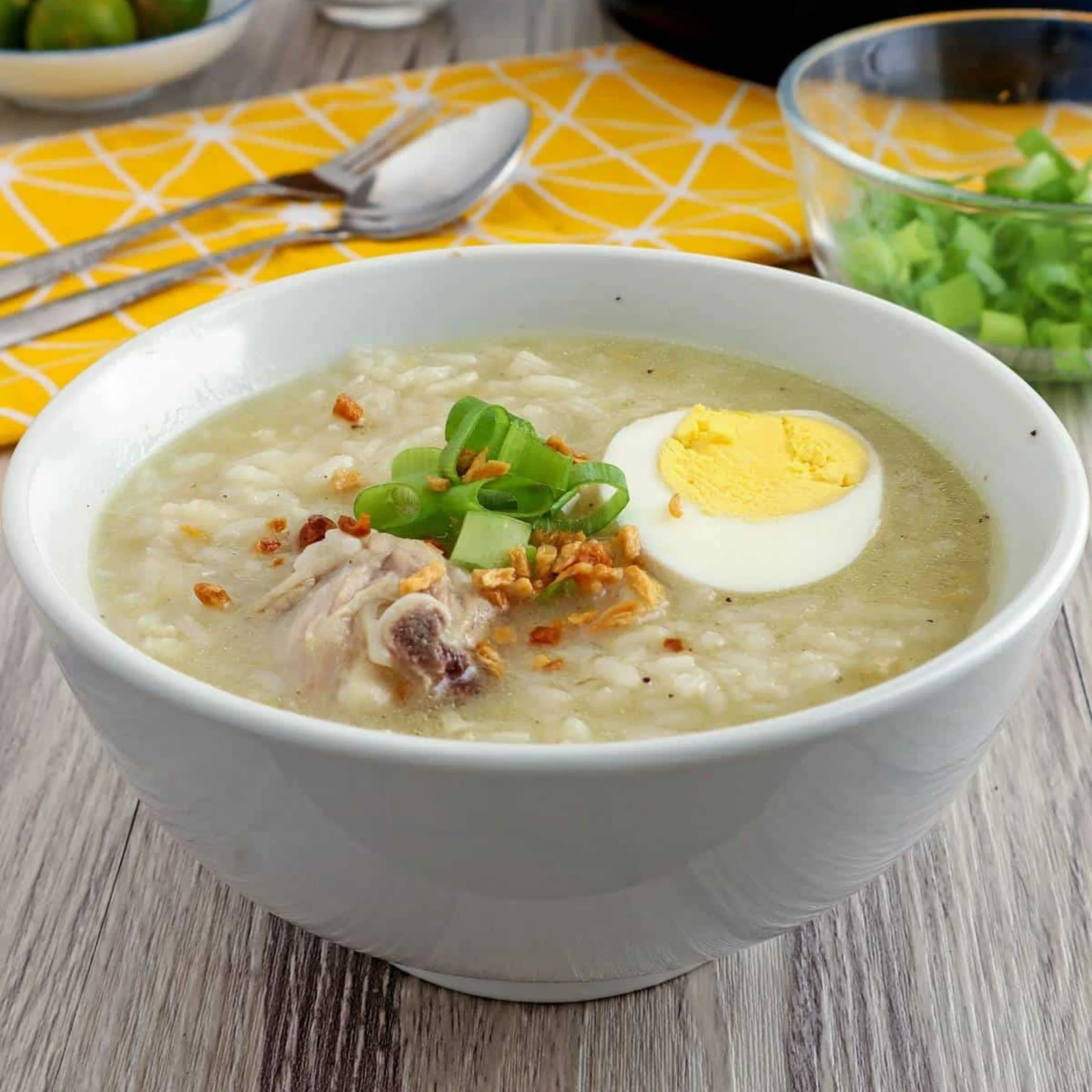 A white bowl of creamy chicken rice porridge, topped with boiled egg, scallions, and fried shallots, sits on a bright yellow tablecloth.