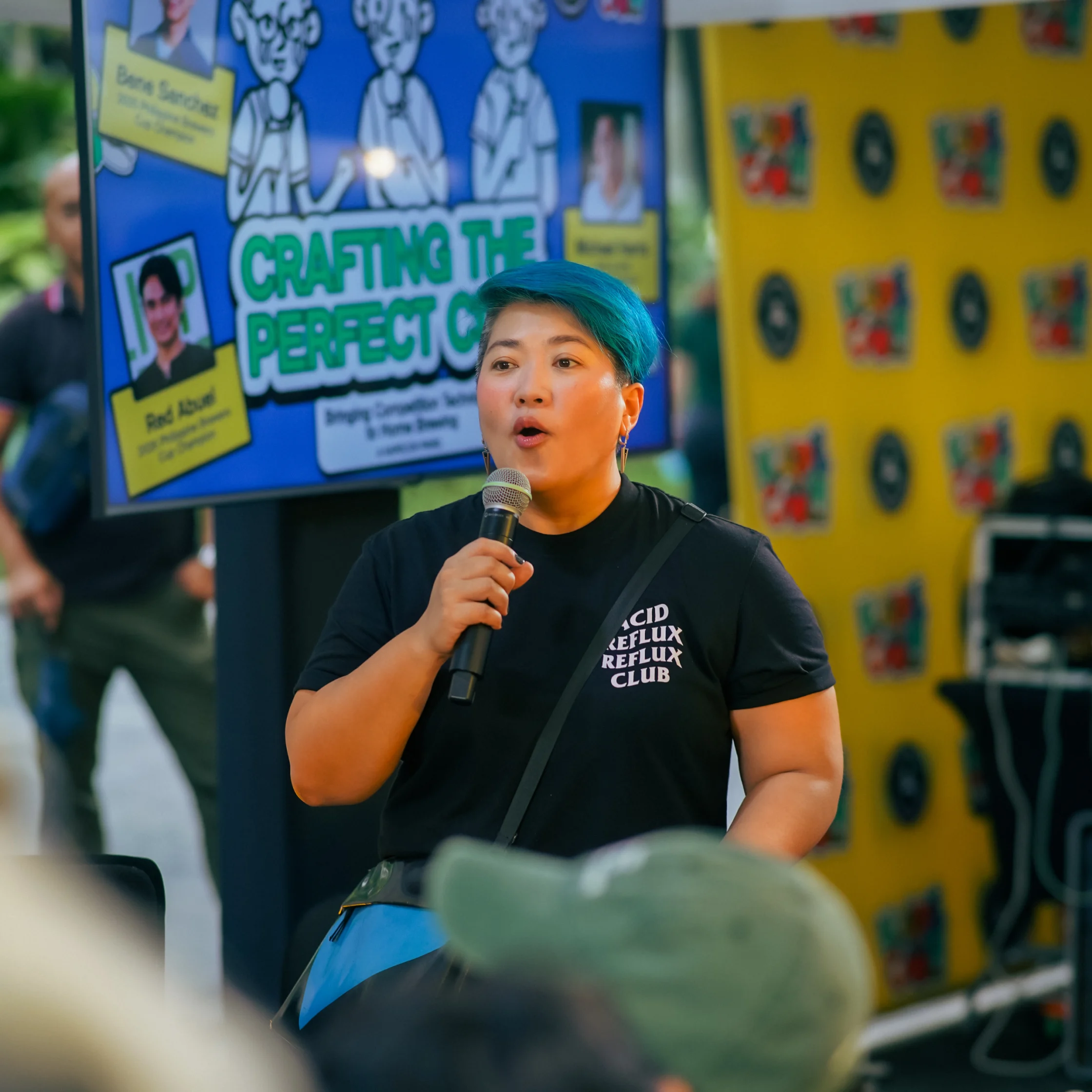 A teal-haired speaker holds a microphone before a colorful cartoon poster and yellow display wall, addressing a crowd in an upbeat, creative event setting.