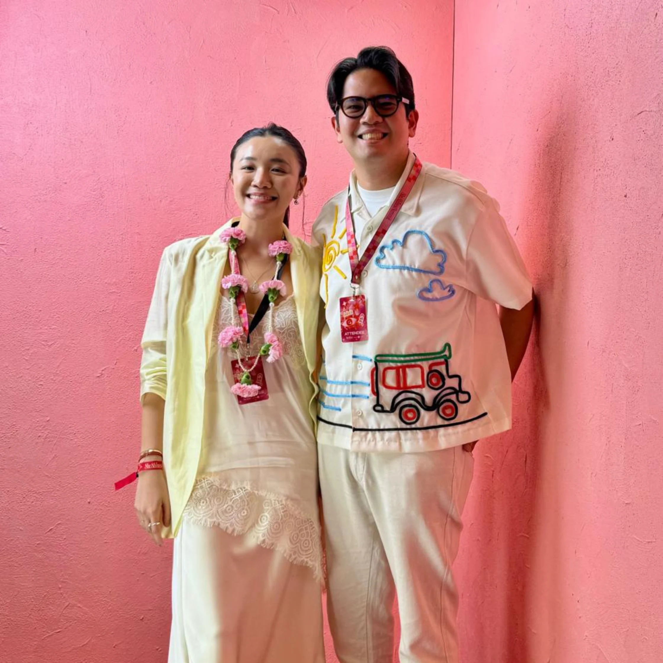 A smiling couple pose before a bright pink wall, wearing light outfits; her floral garland and his playful hand-drawn shirt add a cheerful, festive feel.