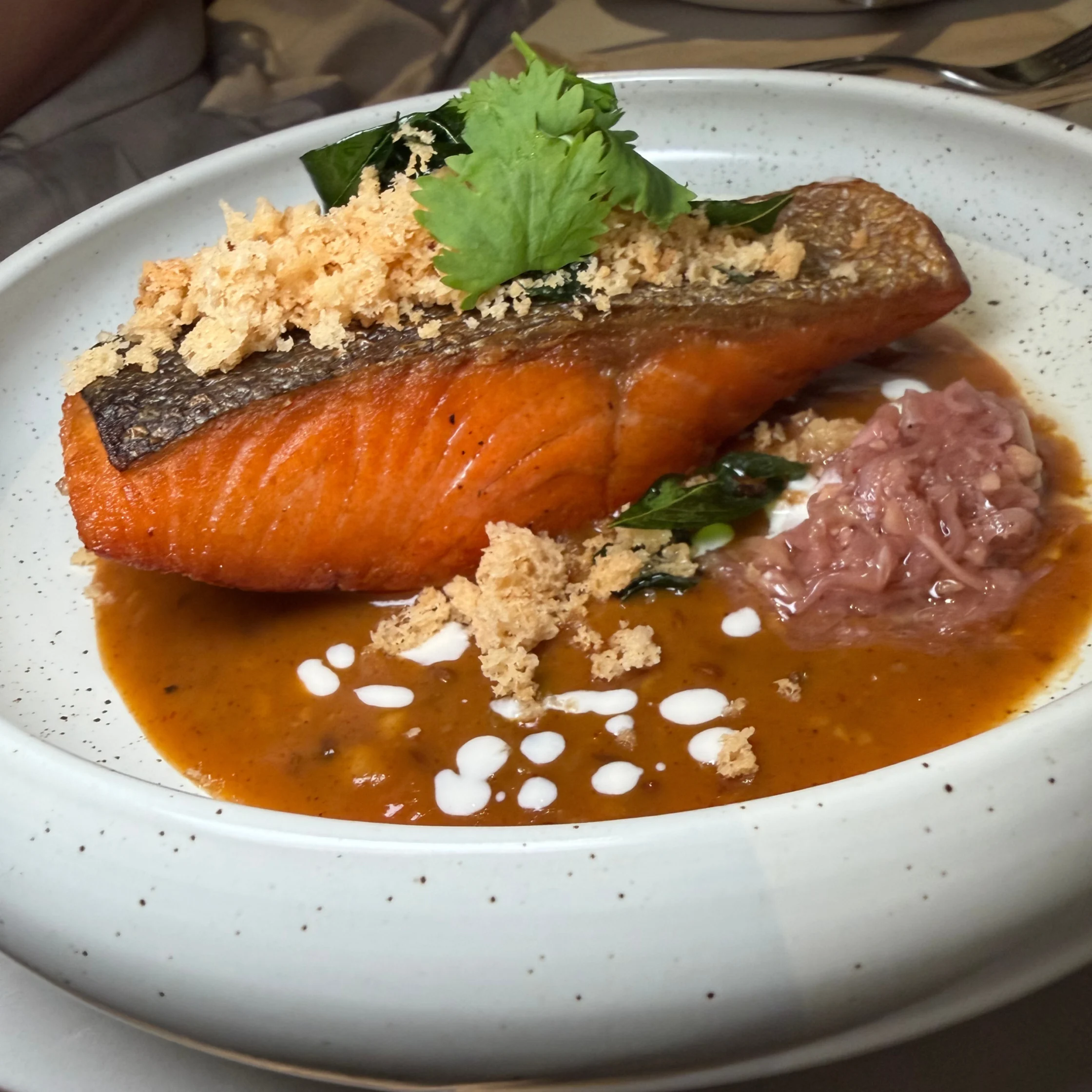 Seared salmon fillet with crisped skin sits in glossy brown sauce, topped with green herbs and crunchy crumbs, with pink relish and white cream dots. A refined, appetizing plated dish.