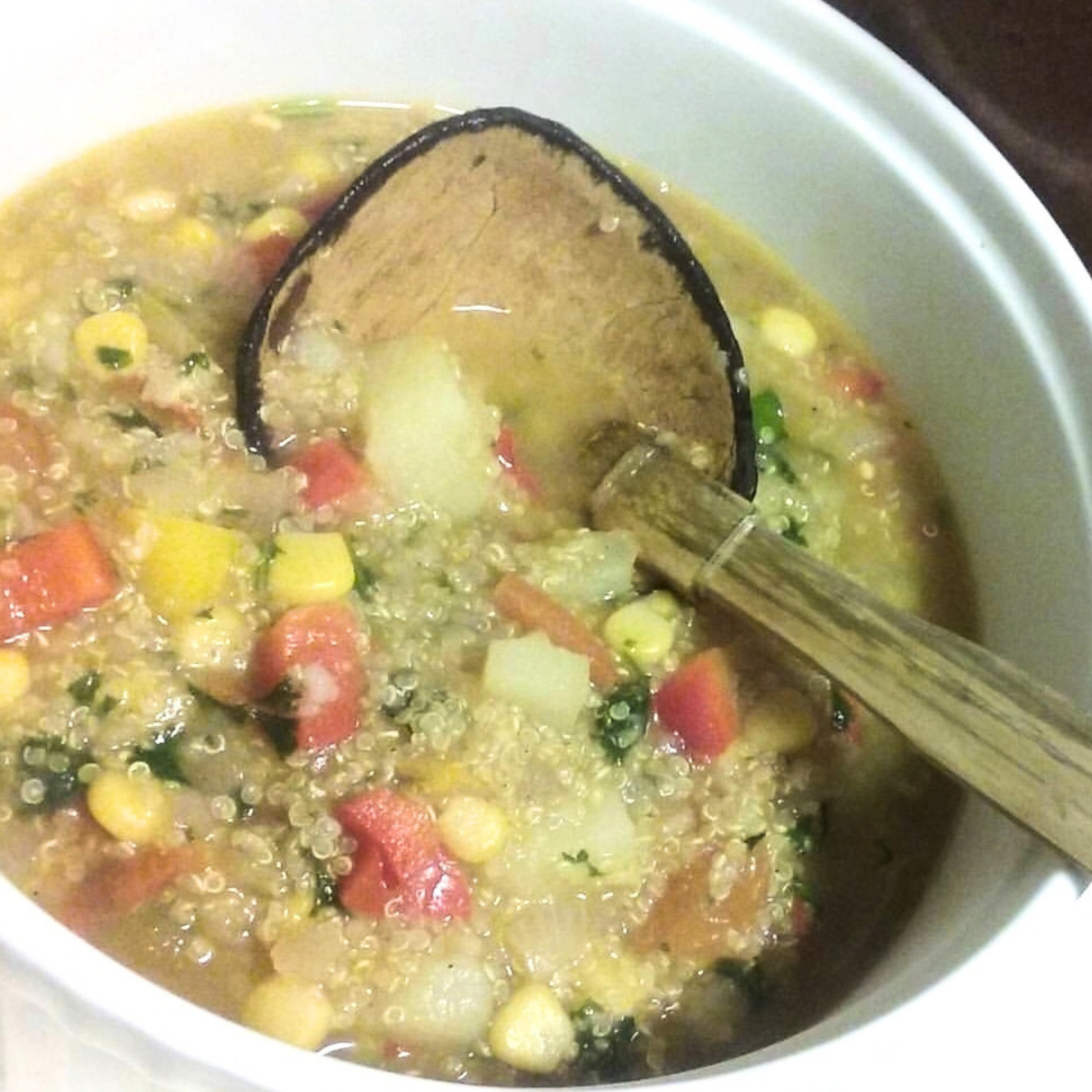 A white pot holds steaming-looking vegetable and grain soup with corn, peas, diced carrots and tomatoes, stirred with a wooden spoon; a hearty, home-cooked meal.
