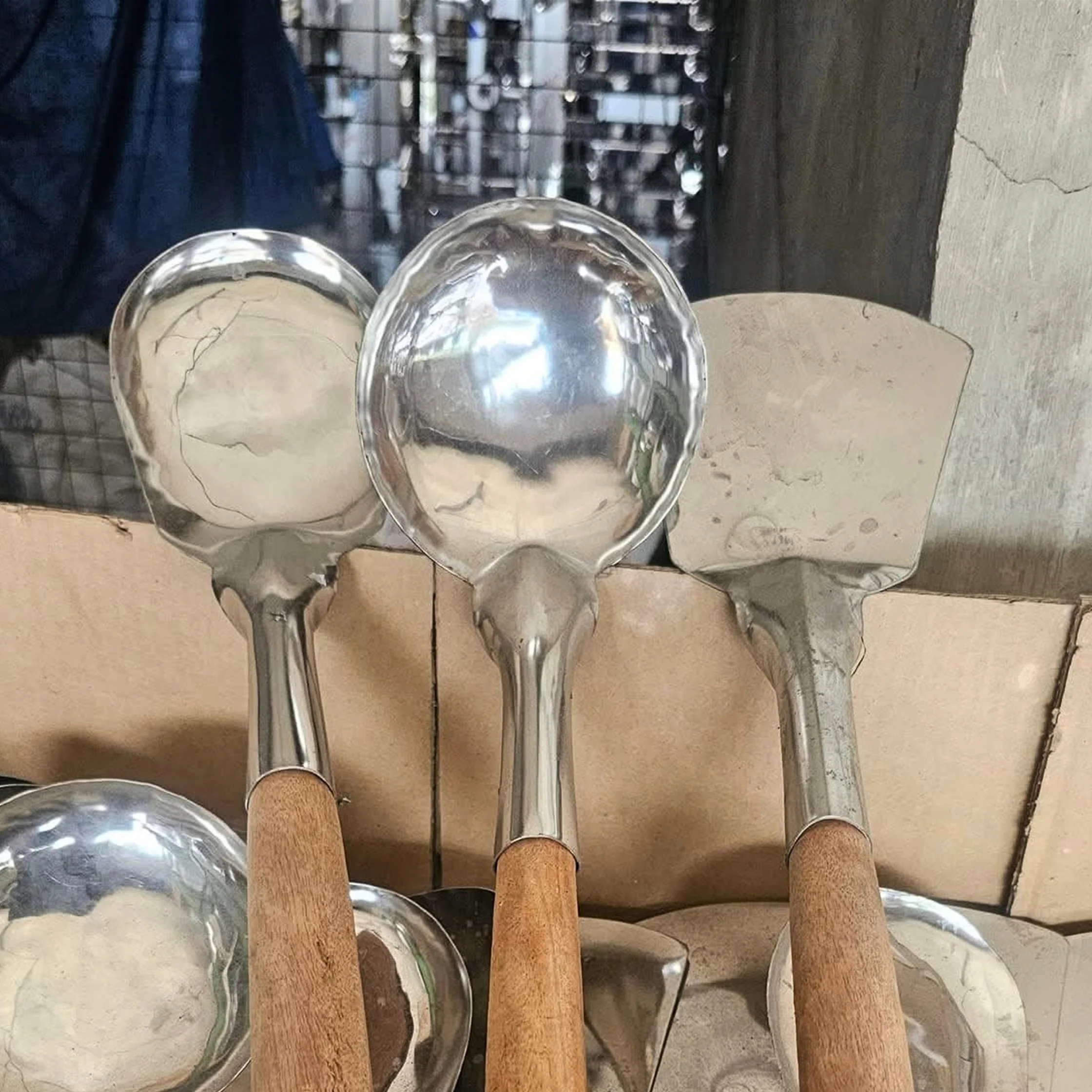 Three worn metal serving utensils with long wooden handles lean against cardboard by a window, their reflective surfaces creating a quiet, utilitarian mood.