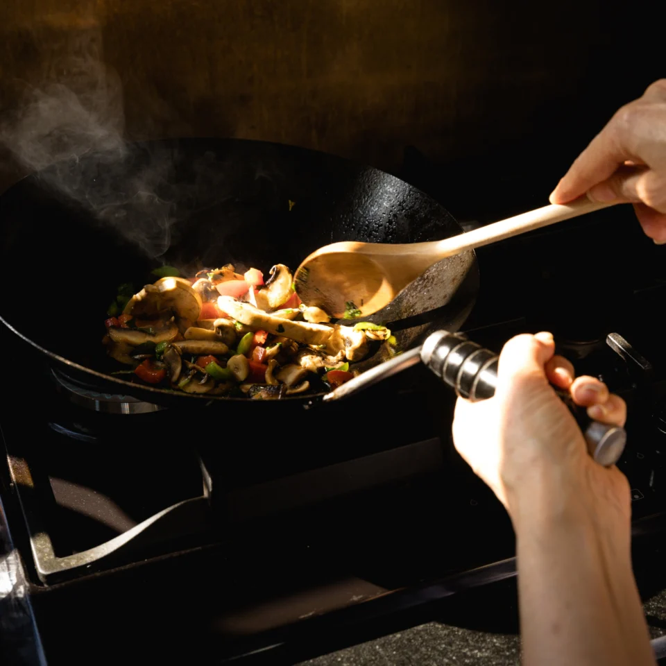 Hands stir sizzling vegetables in a steaming black wok on a stovetop, using a wooden spoon and metal-handled utensil; warm light creates a cozy cooking scene.