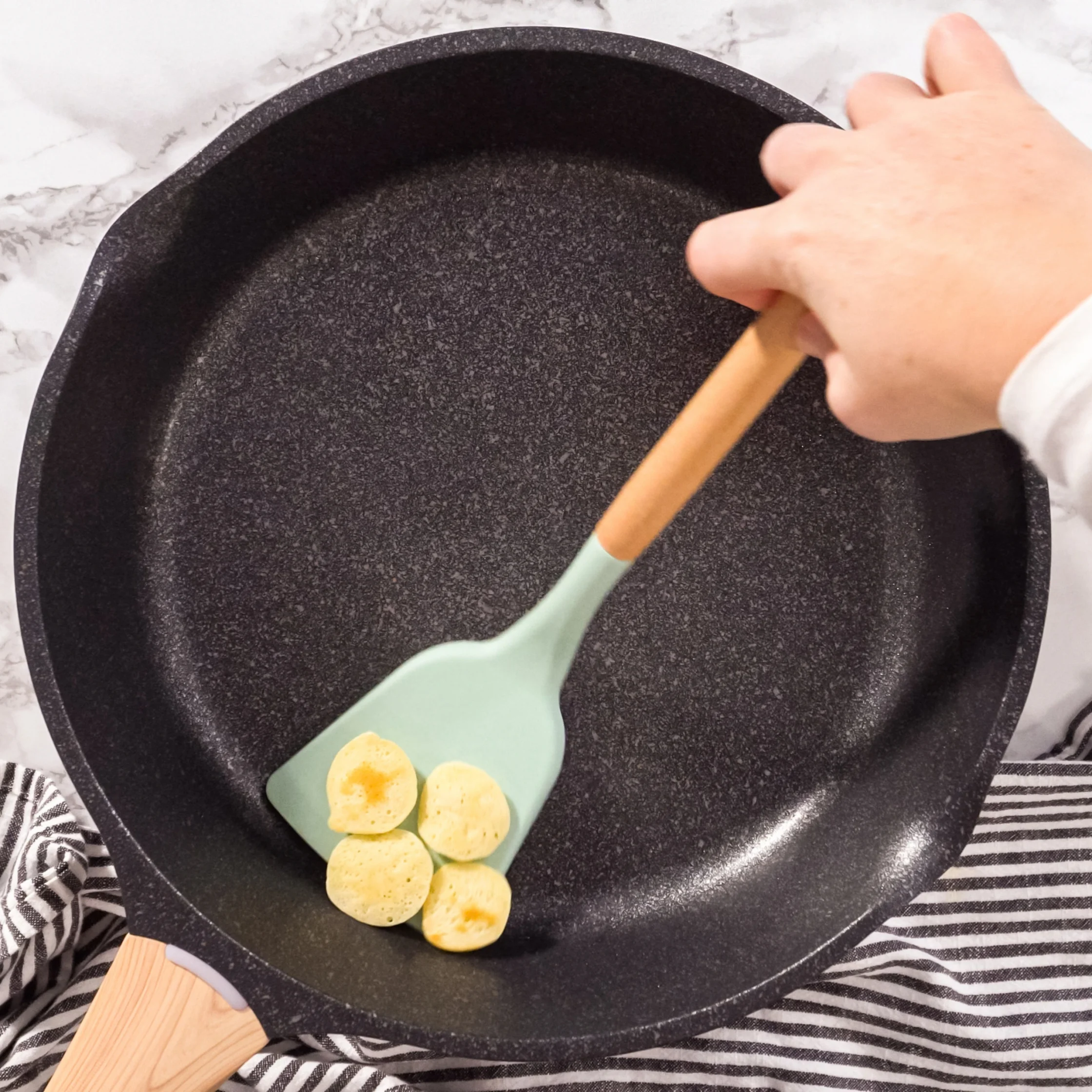 A hand uses a mint-green spatula to turn four small pancakes in a black frying pan on a marble counter, creating a calm, homey cooking scene.