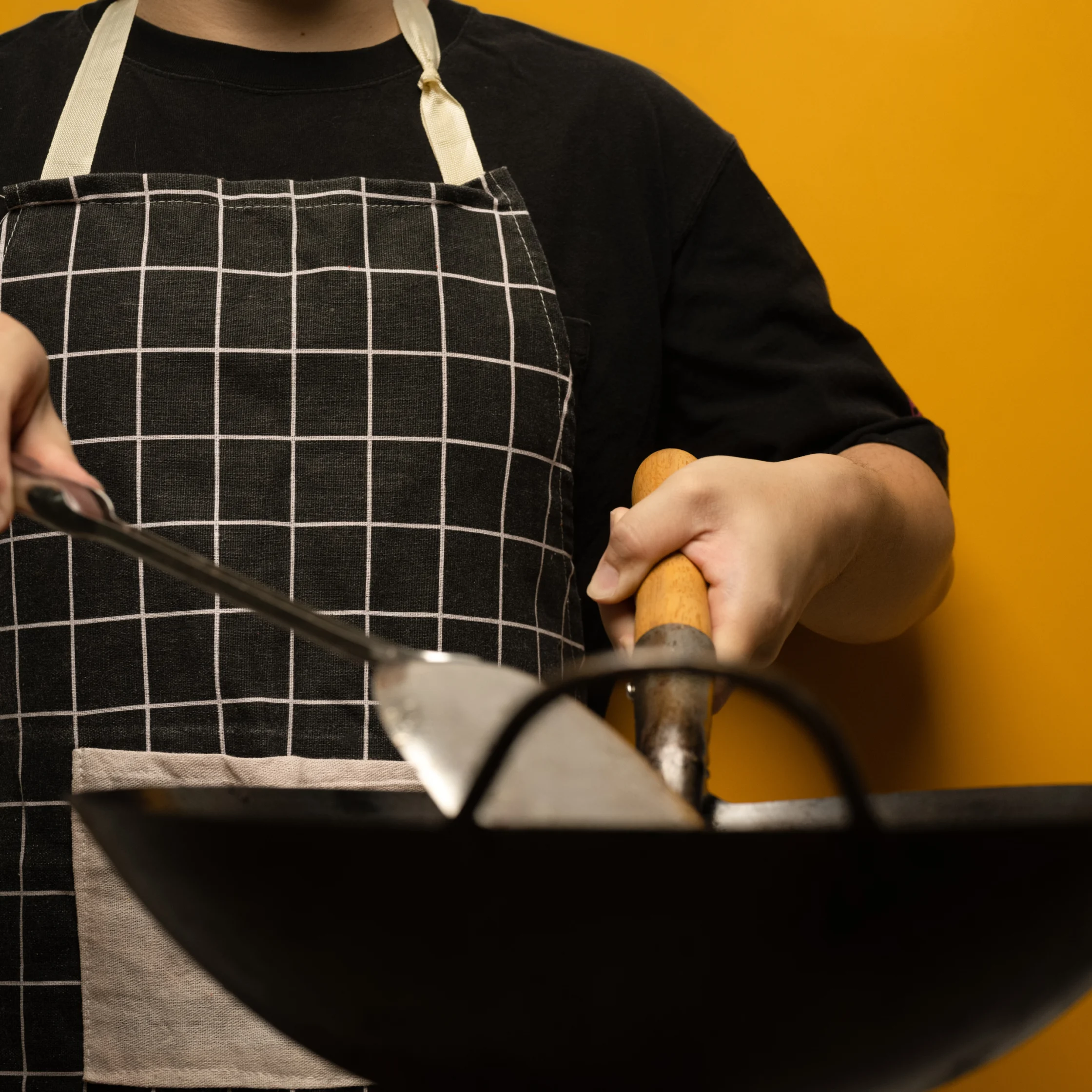 A cook in a black shirt and checked apron stirs a steaming wok with a metal spatula, gripping its wooden handle against a warm yellow background.