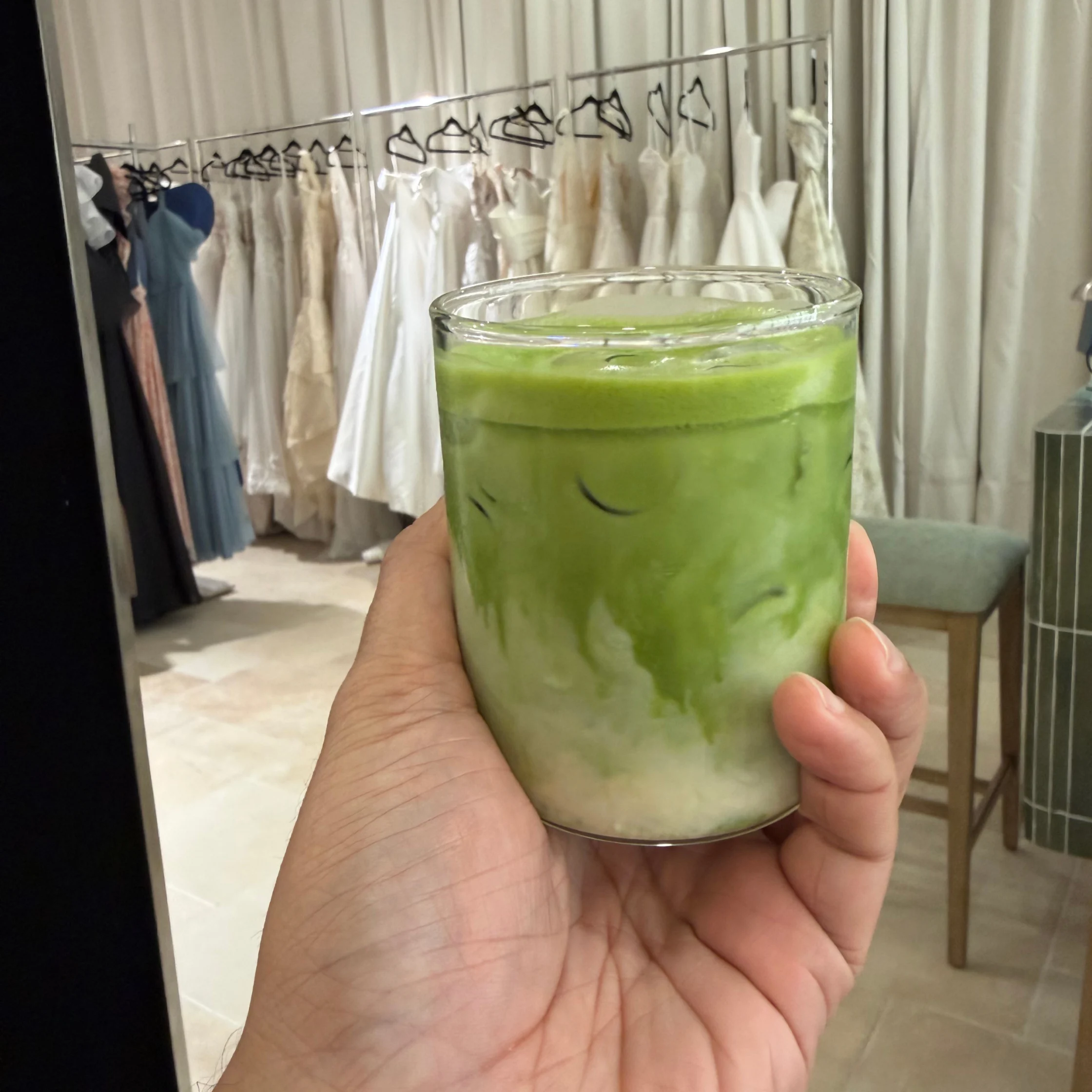 A hand holds a glass of iced matcha latte, its green layers swirling with milk, in a calm boutique clothing shop with dresses on a rack.