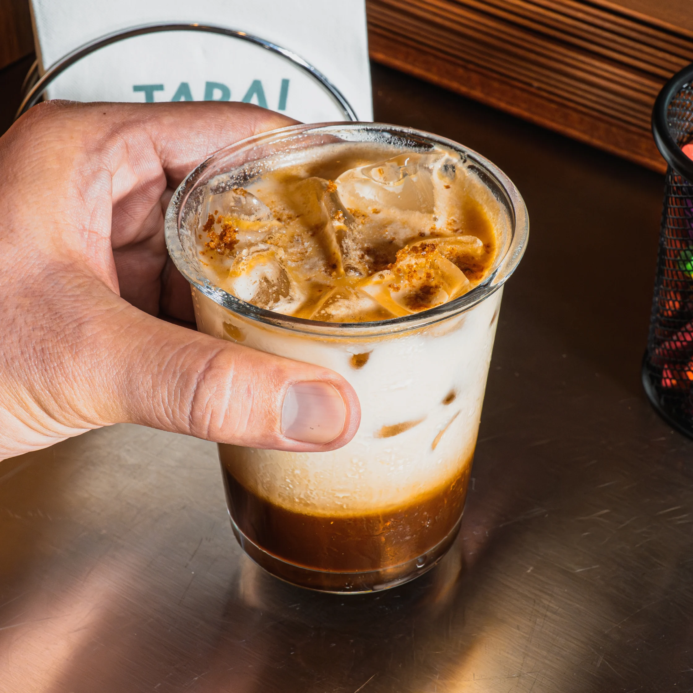 A hand holds a clear cup of iced coffee layered with dark espresso, milk, and ice on a café counter, conveying a casual, refreshing mood.