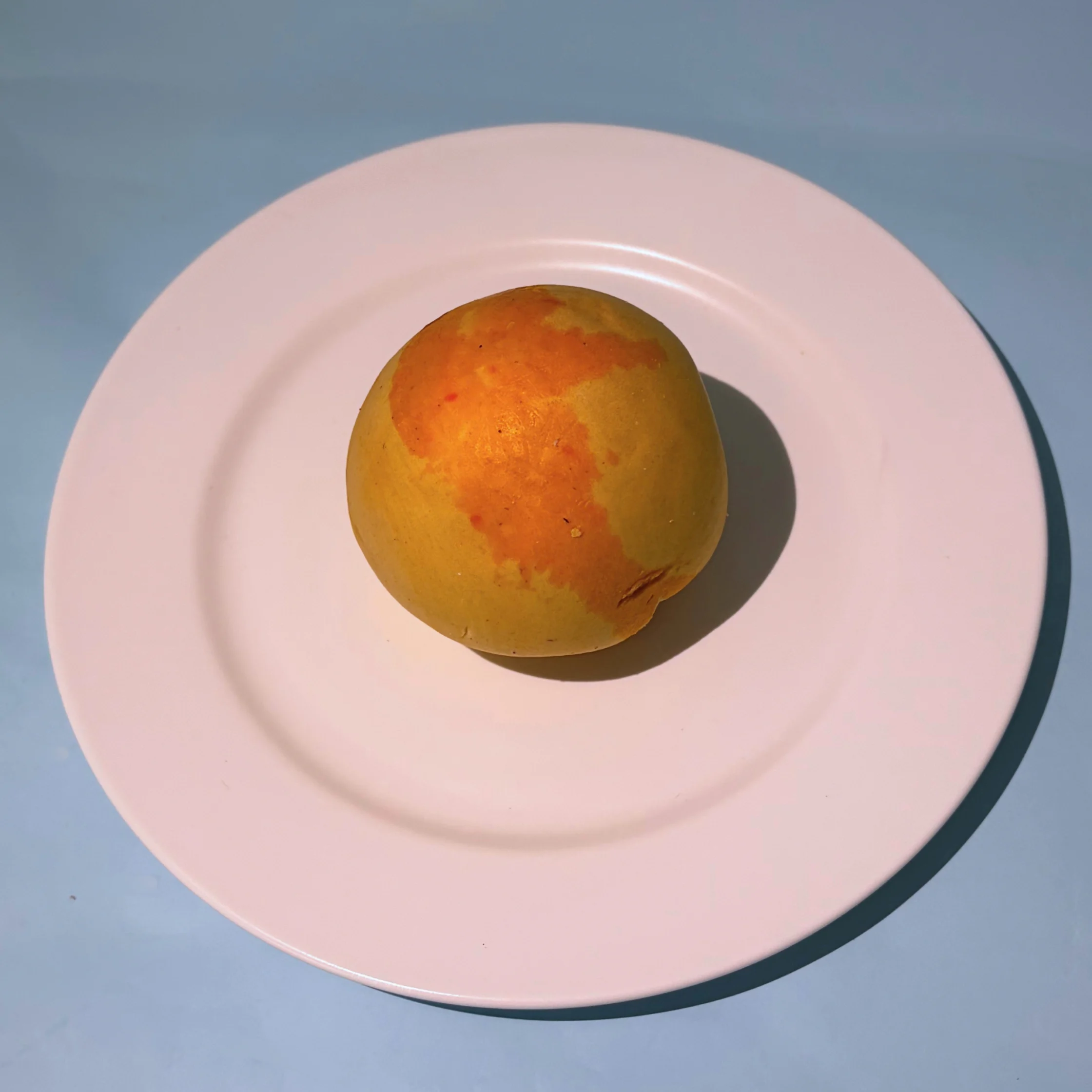 A ripe yellow-orange mango sits alone on a pale pink plate against a blue-gray surface, casting a sharp shadow in bright light.