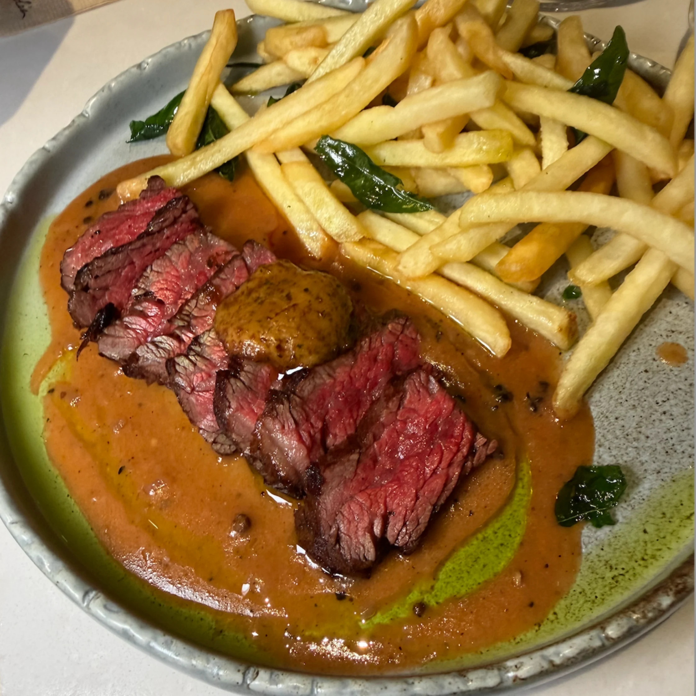 Sliced medium-rare steak rests in rich brown sauce beside golden fries and green herbs on a ceramic plate, creating a hearty, appetizing meal.