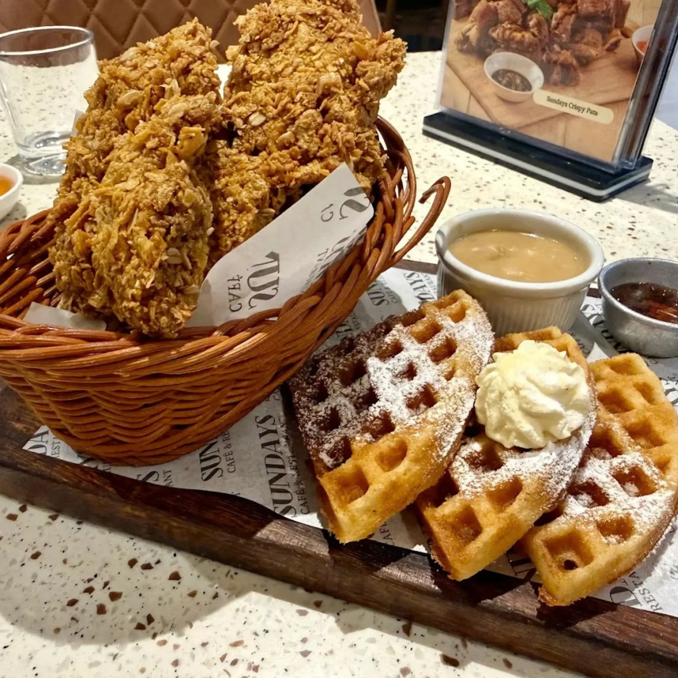 A basket piled with golden fried chicken sits beside waffles dusted with powdered sugar and topped with cream, with dipping sauces on a restaurant table.