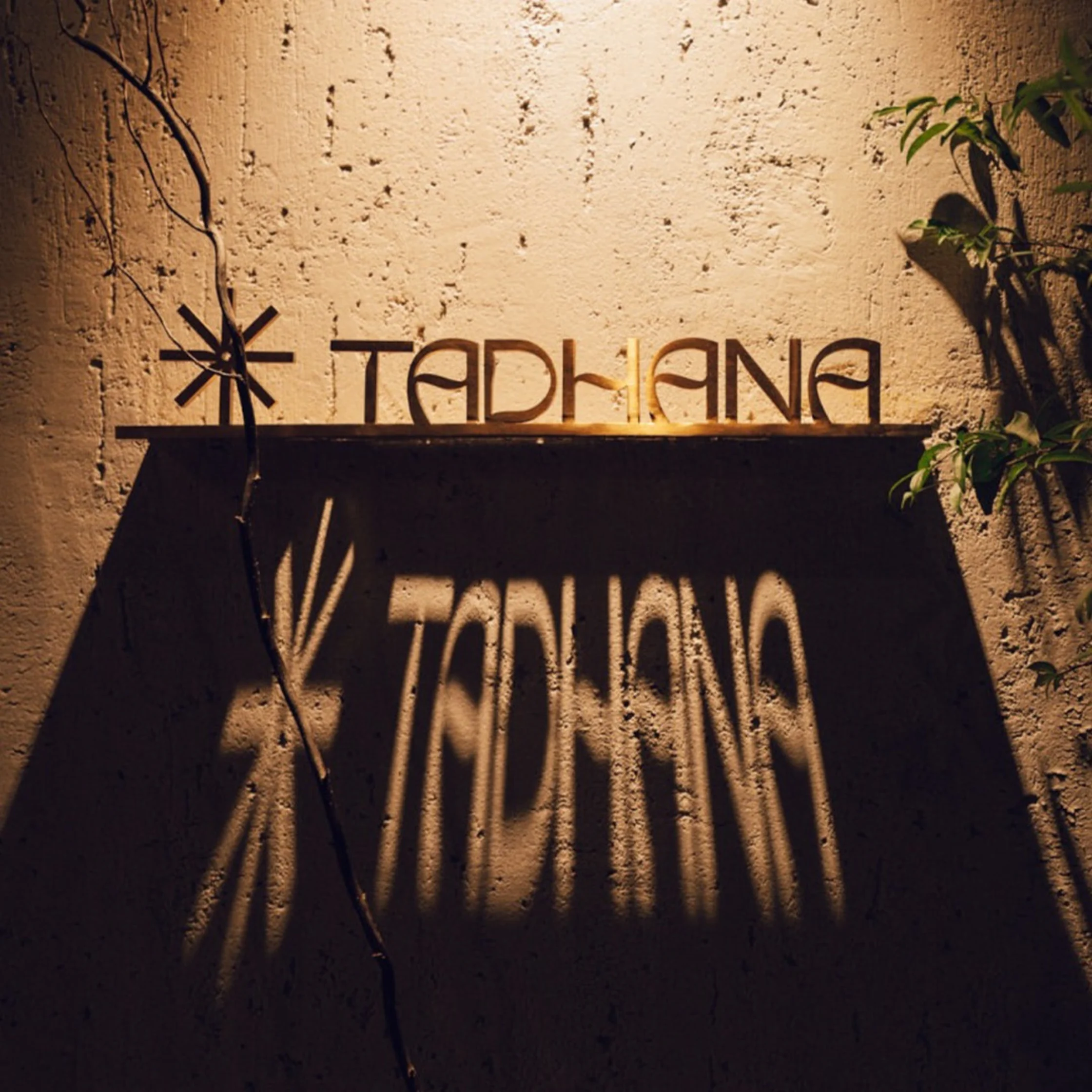 A rustic tan stucco wall bears a metal “TADHANA” sign, casting long, dramatic shadows beside sparse greenery in warm afternoon light.