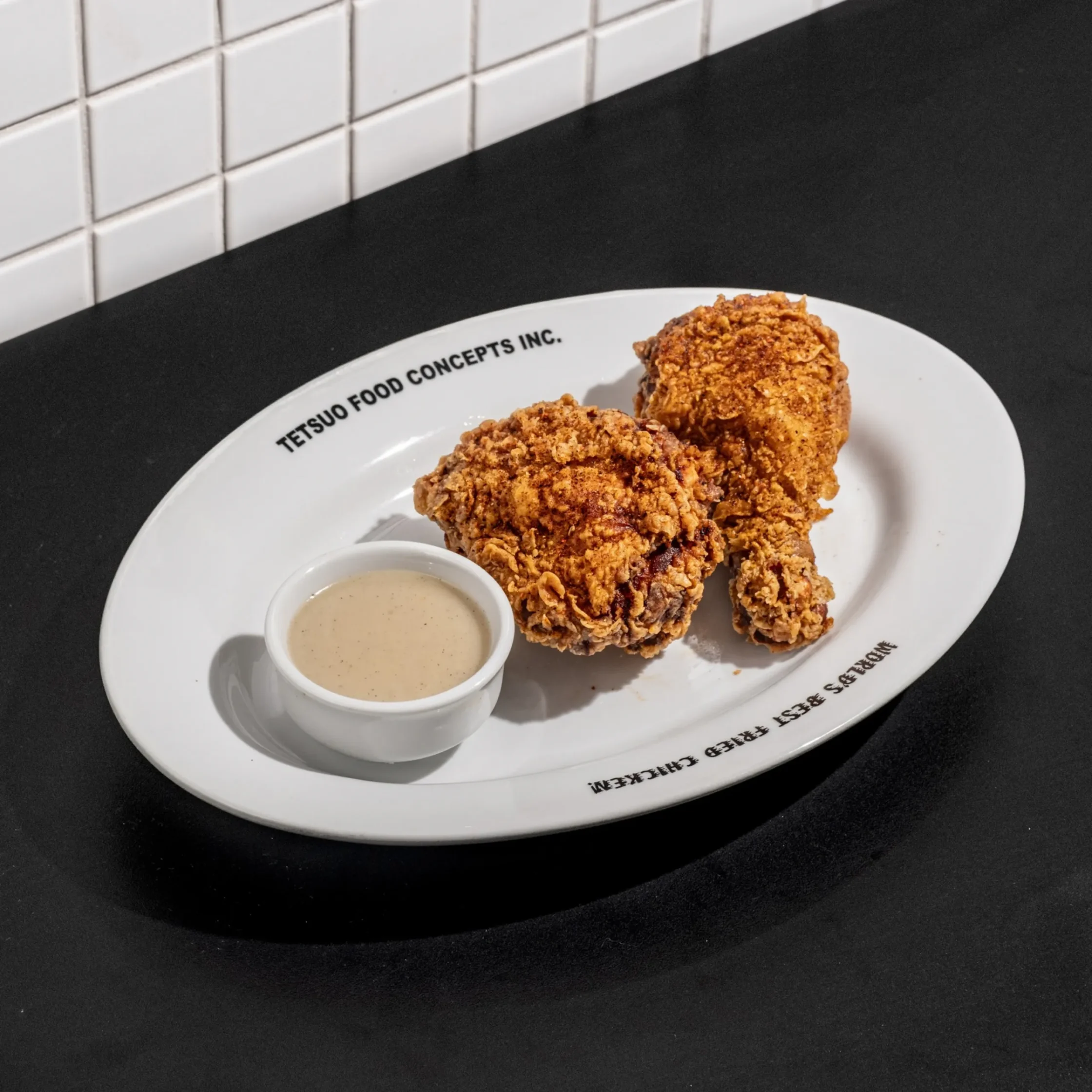 Two pieces of golden, crispy fried chicken and a small bowl of creamy dipping sauce sit on a white oval plate against a black countertop.