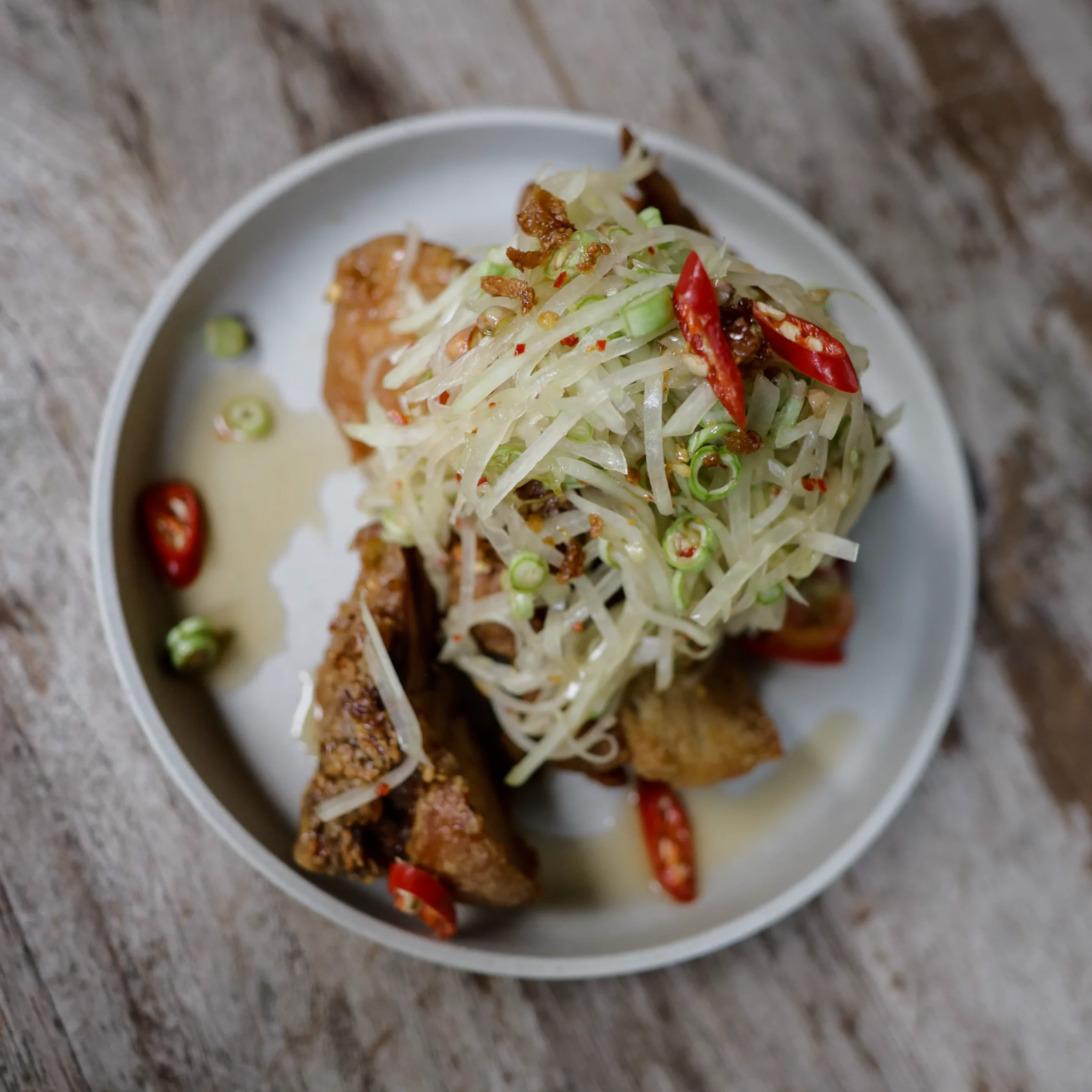 Top-down view of crispy browned meat topped with shredded green papaya salad, red chilies, scallions, and herbs on a gray plate; rustic, appetizing meal.