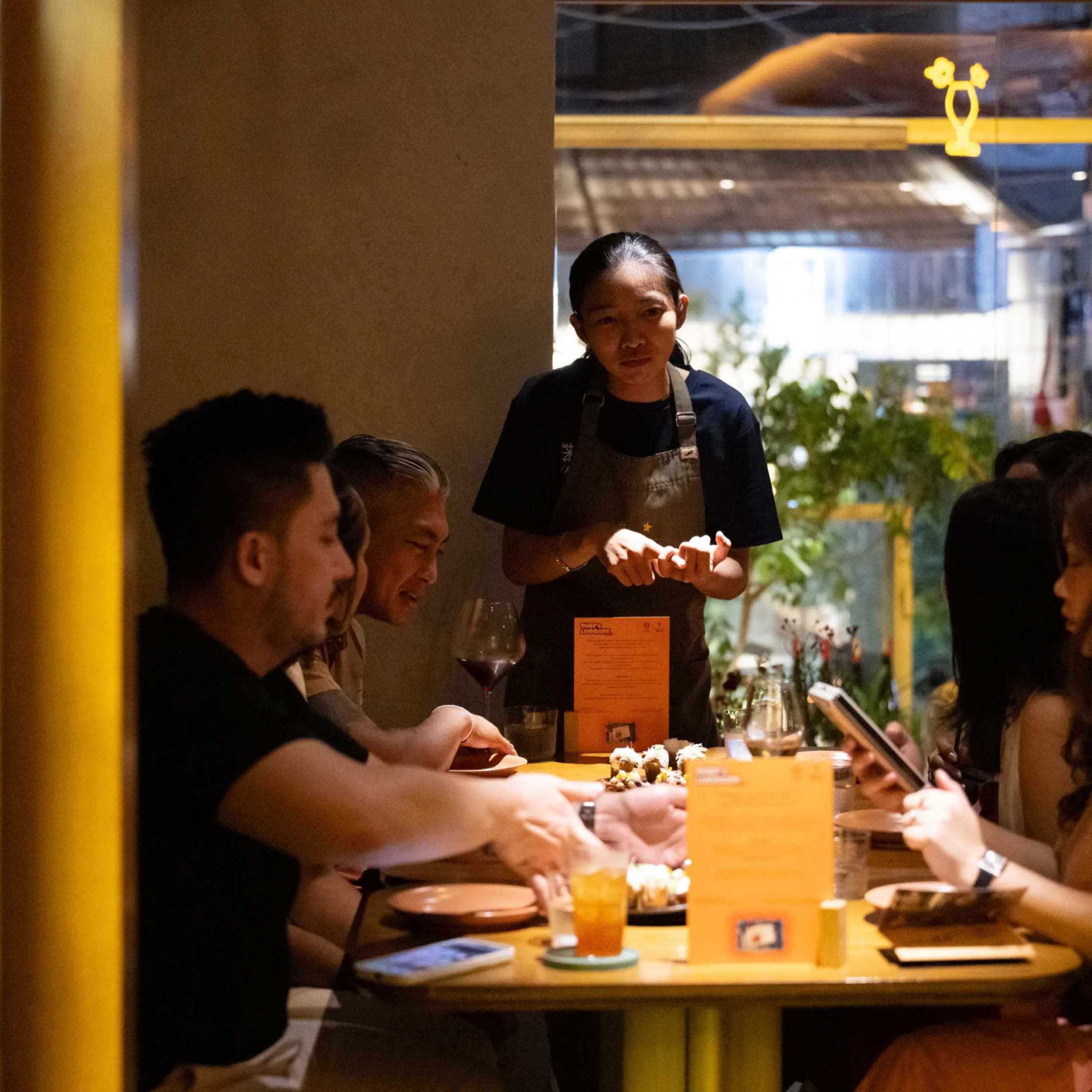 A server explains a dish to diners gathered around a warmly lit restaurant table, where plates, drinks, and menus create an intimate, lively scene.