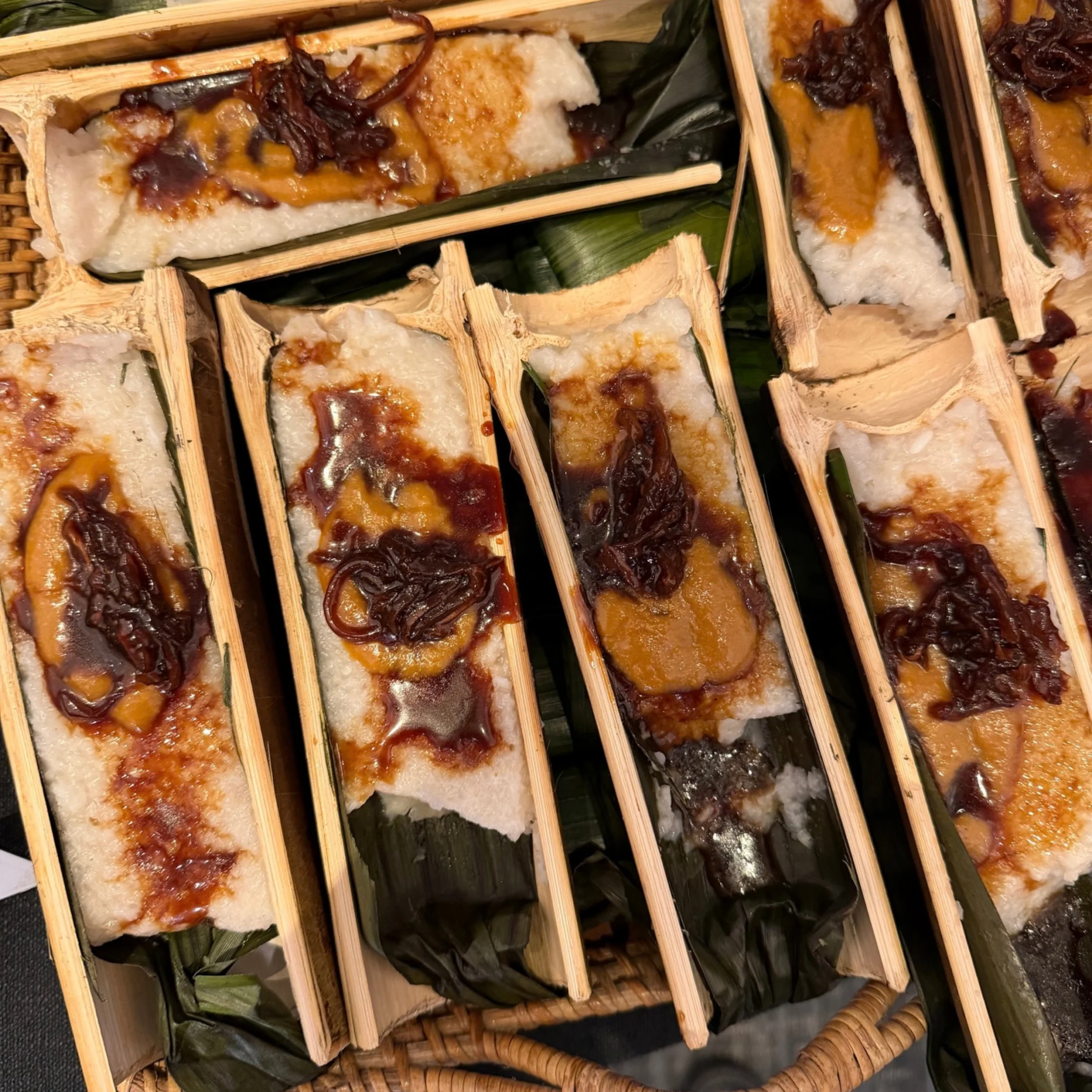 Close-up of sticky rice cakes topped with glossy brown sauce and dark shredded garnish, arranged in split bamboo trays over banana leaves; rustic and appetizing.
