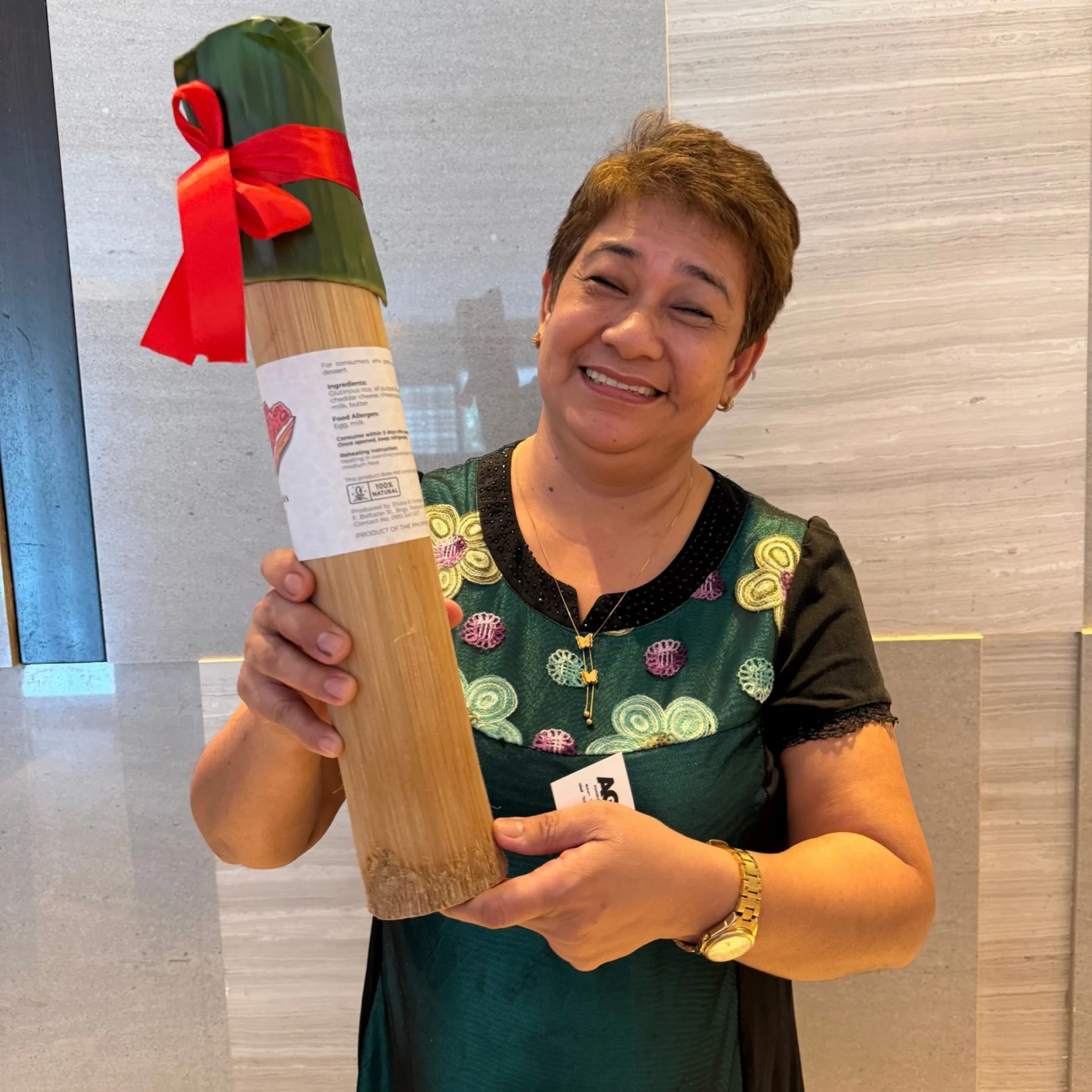 Smiling woman in an embroidered green dress proudly holds a long wrapped food package tied with a bright red ribbon in a modern room.