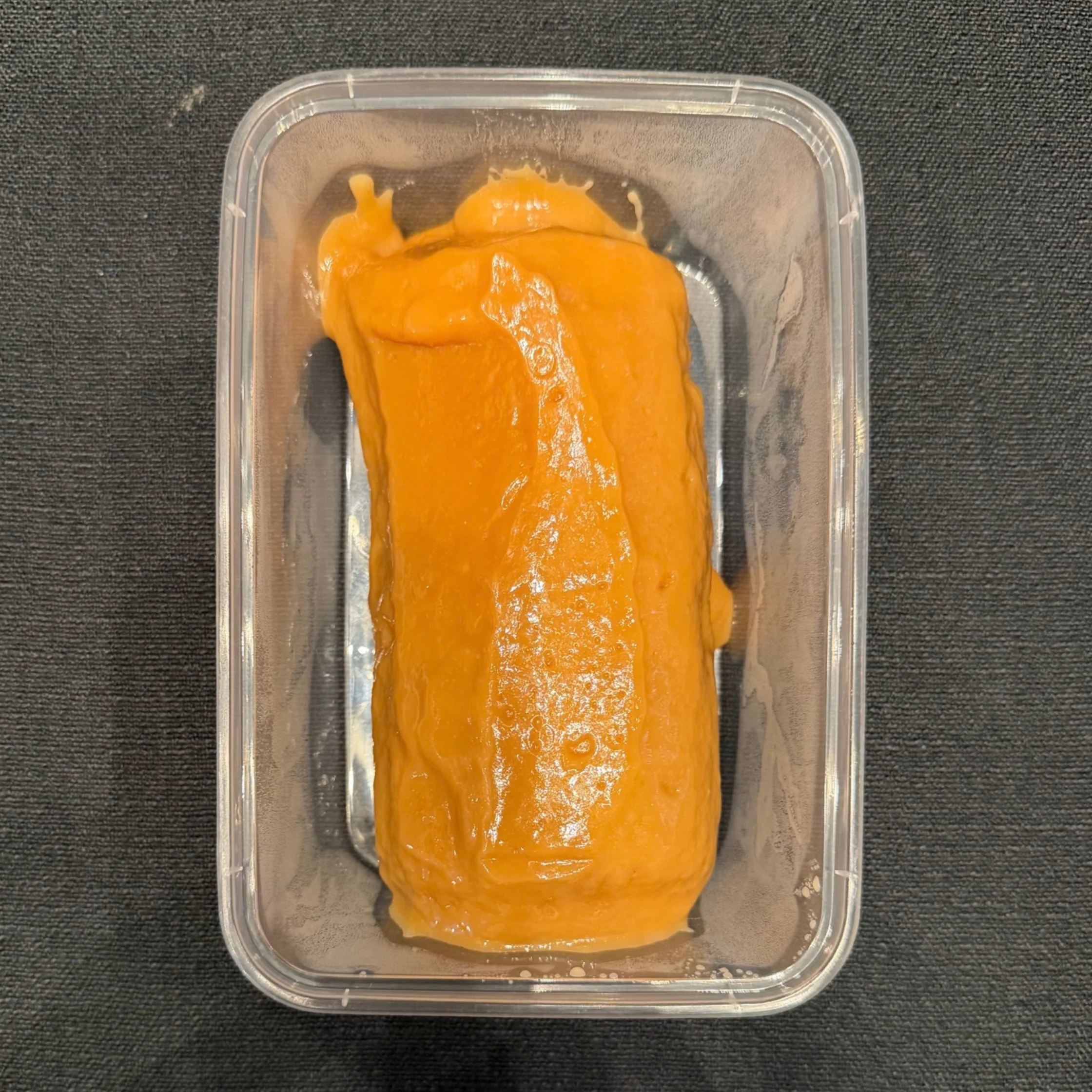 Bright orange, smooth pâté or spread fills a clear rectangular plastic container on a dark fabric surface, with a glossy, homemade appearance.