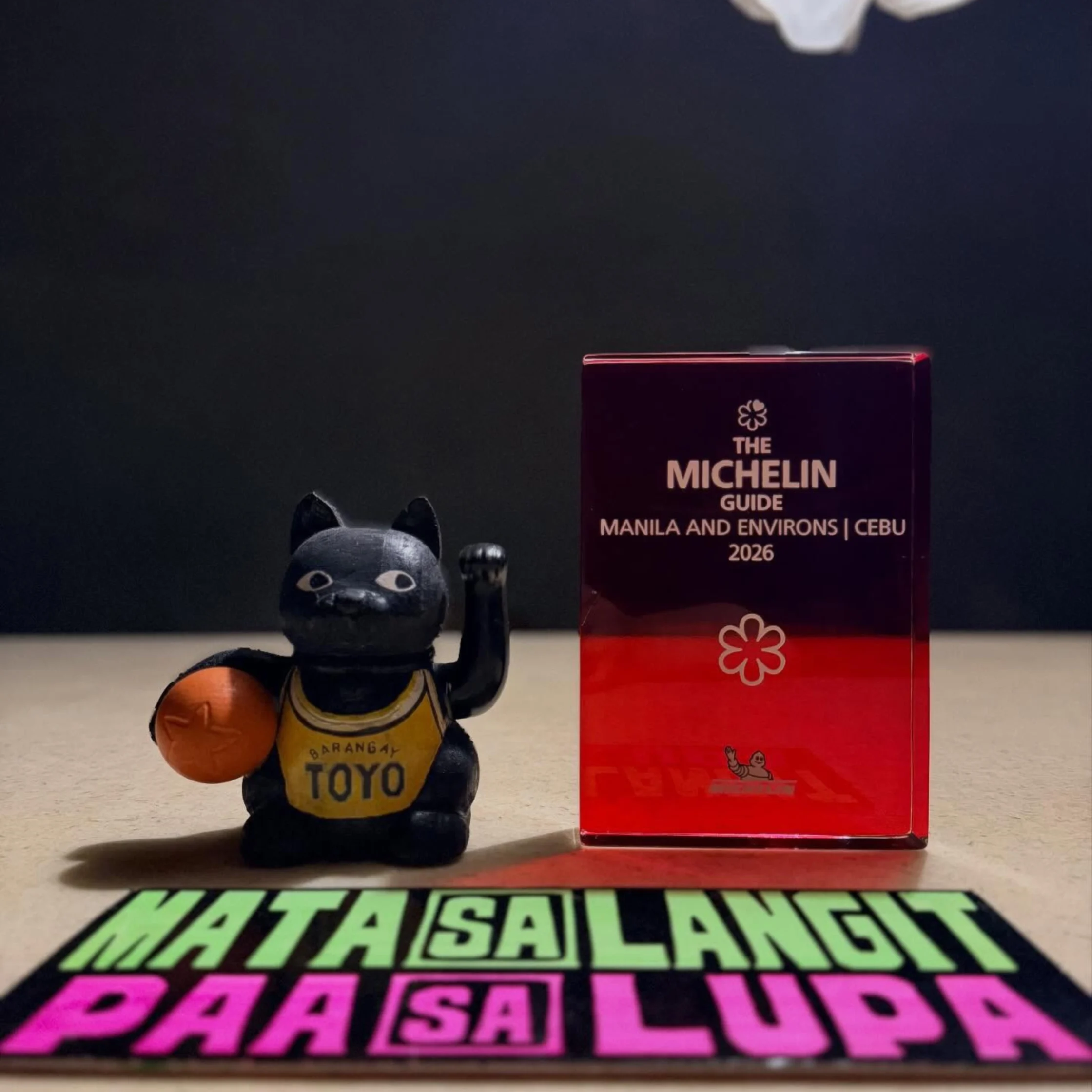A black Maneki-neko cat figurine in a “Toyo” jersey holds an orange ball beside a red Michelin Guide Manila and Environs | Cebu 2026 book, with a bright Tagalog slogan below. The playful display feels celebratory.