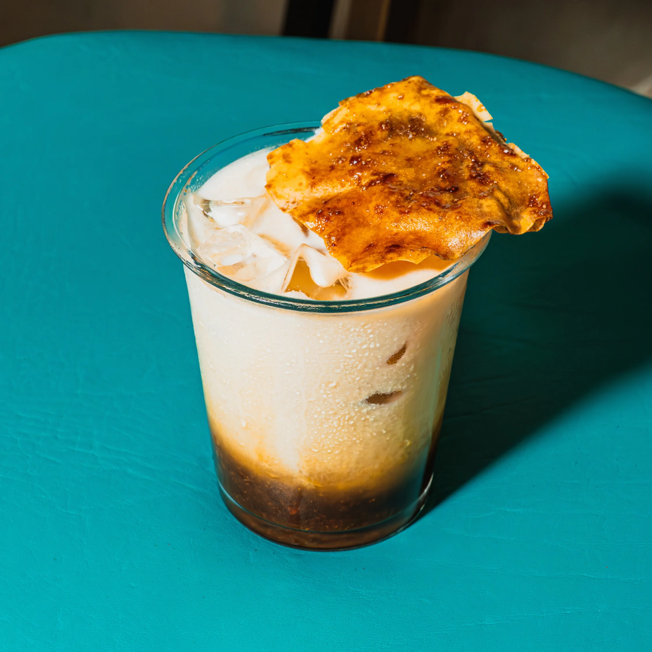 A creamy iced coffee drink with ice in a clear glass, topped with a golden-brown toasted pastry, sits on a bright teal table. The scene feels inviting.