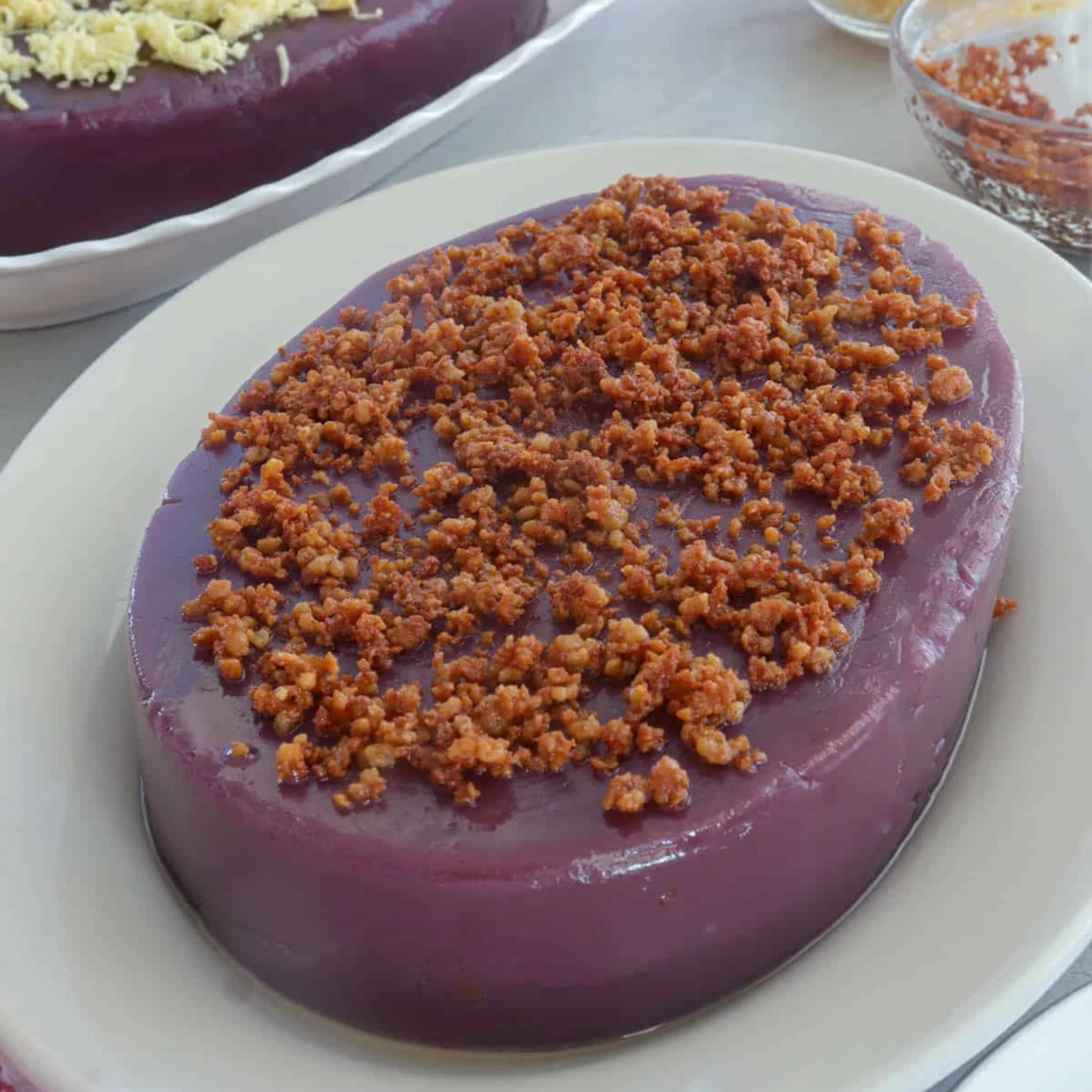 A smooth purple dessert on a white oval platter is topped with a generous layer of golden-brown crumbs; more desserts sit nearby, creating an inviting spread.