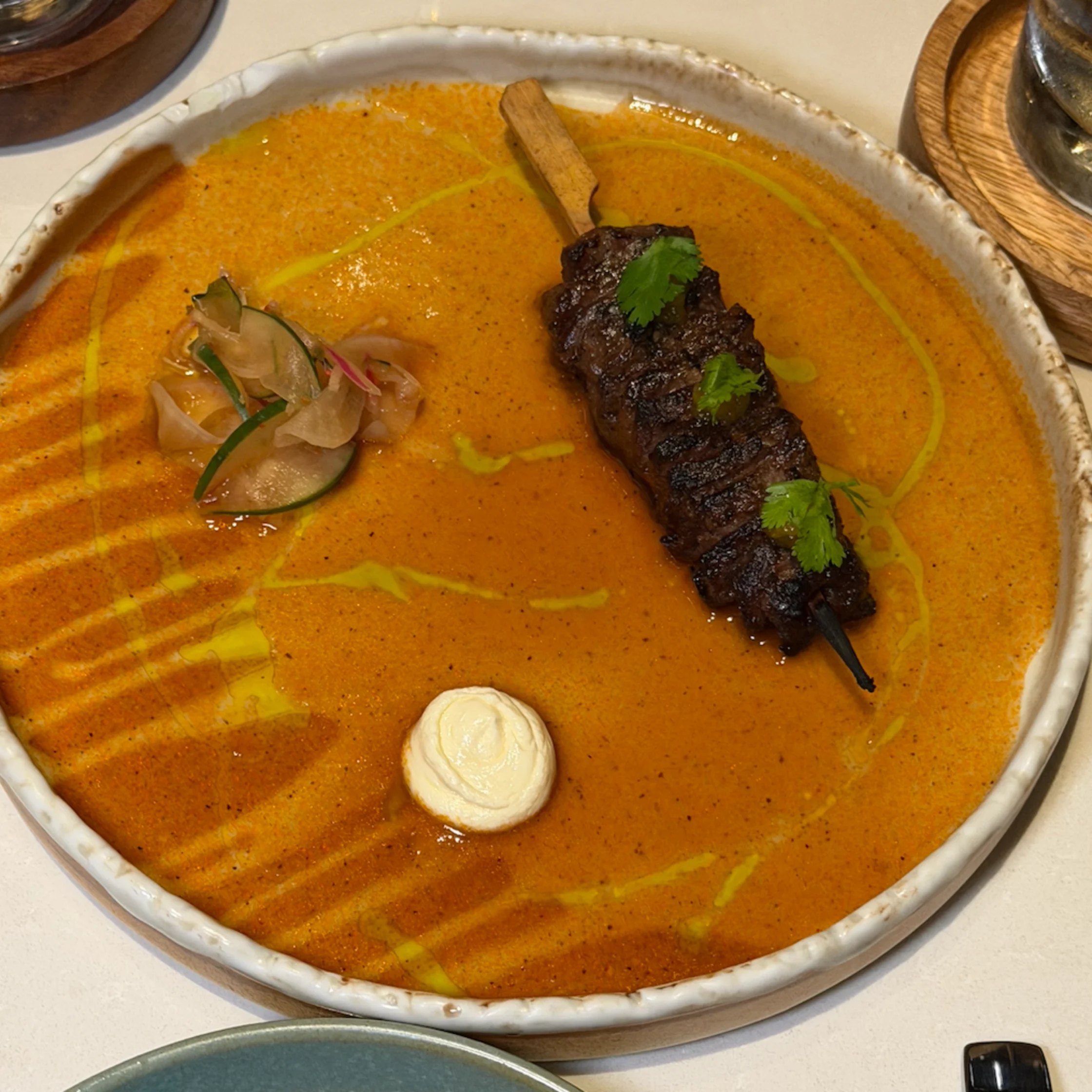 A grilled meat kebab garnished with cilantro rests in a bowl of orange curry, drizzled with green oil and served with pickled onions and cream.